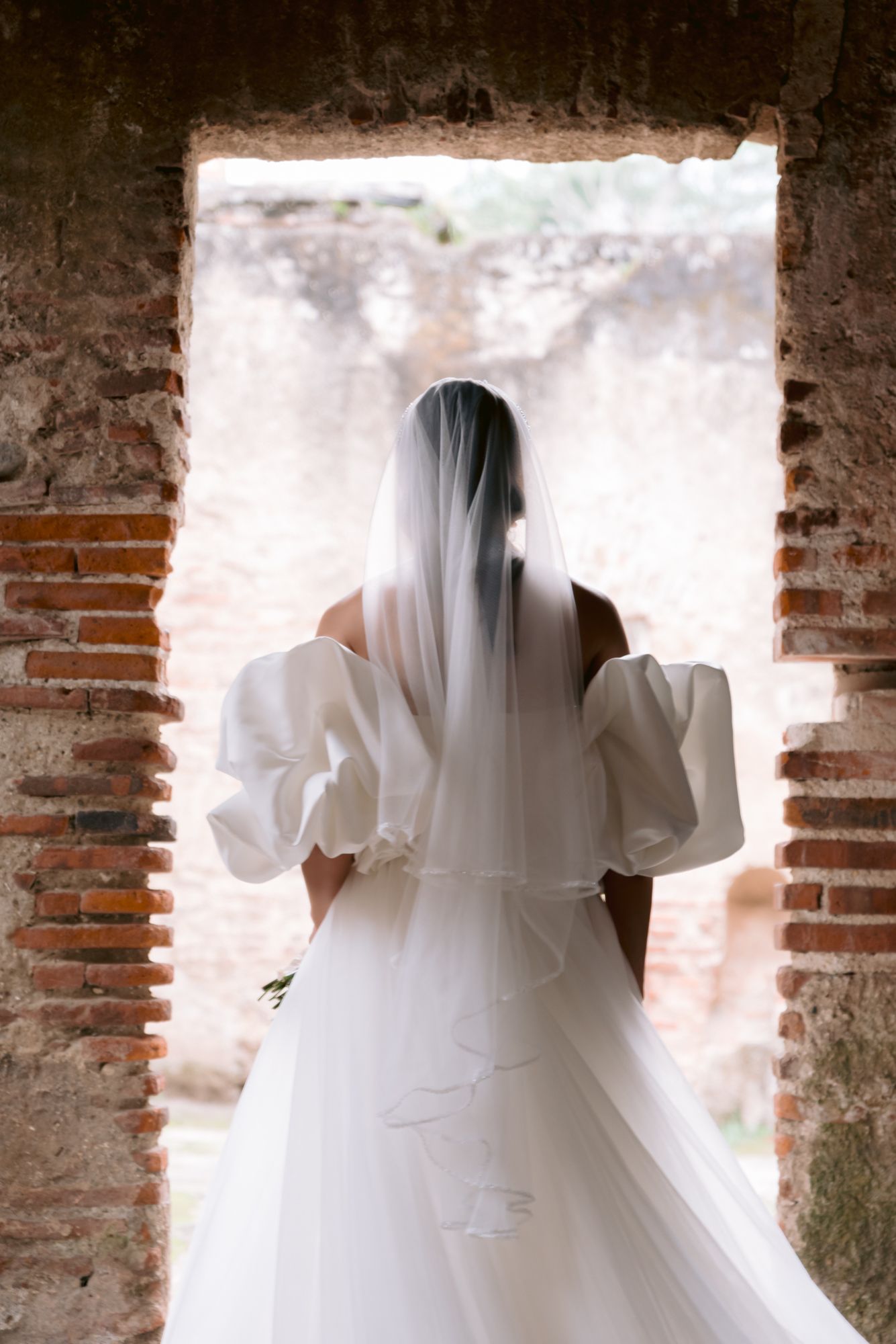 Silhouette of bride in puff sleeve wedding dress framed by sunlit brick archway.