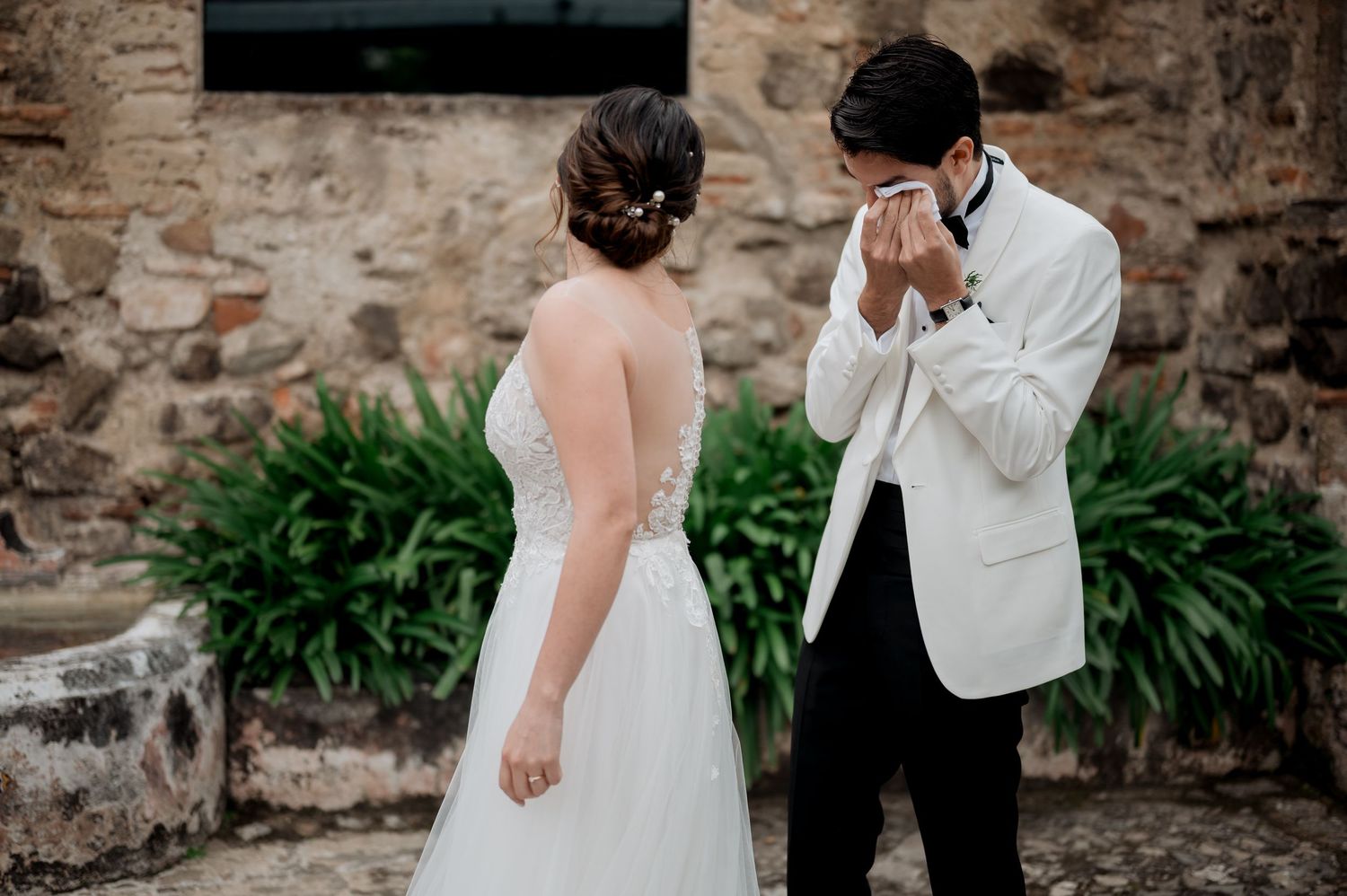 Candid wedding reaction against textured stone wall and green plants.