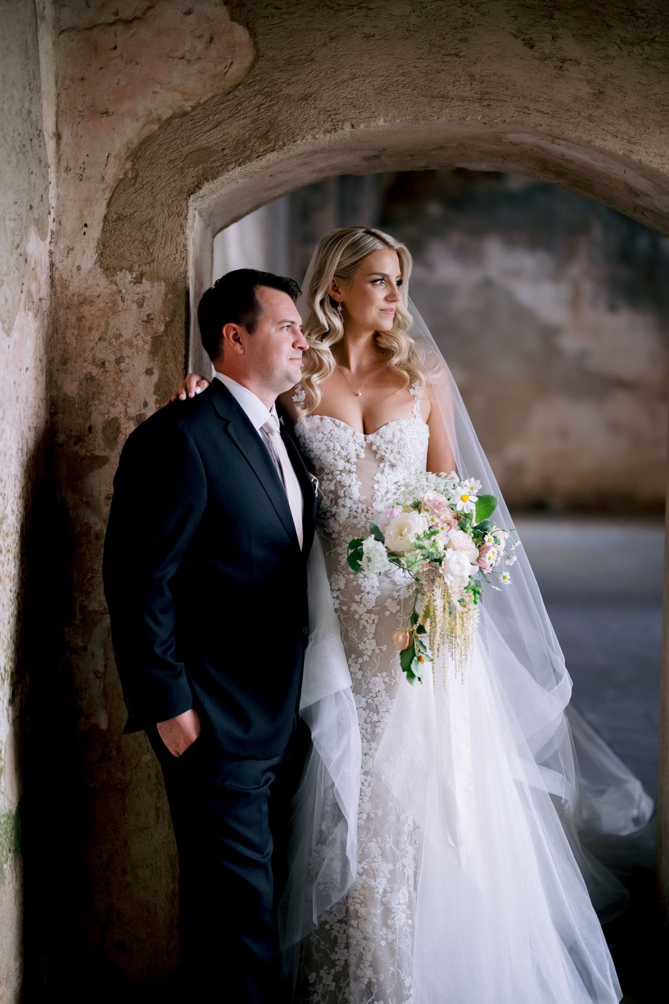 Wedding couple poses in historic stone archway with elegant bouquet and formal attire.