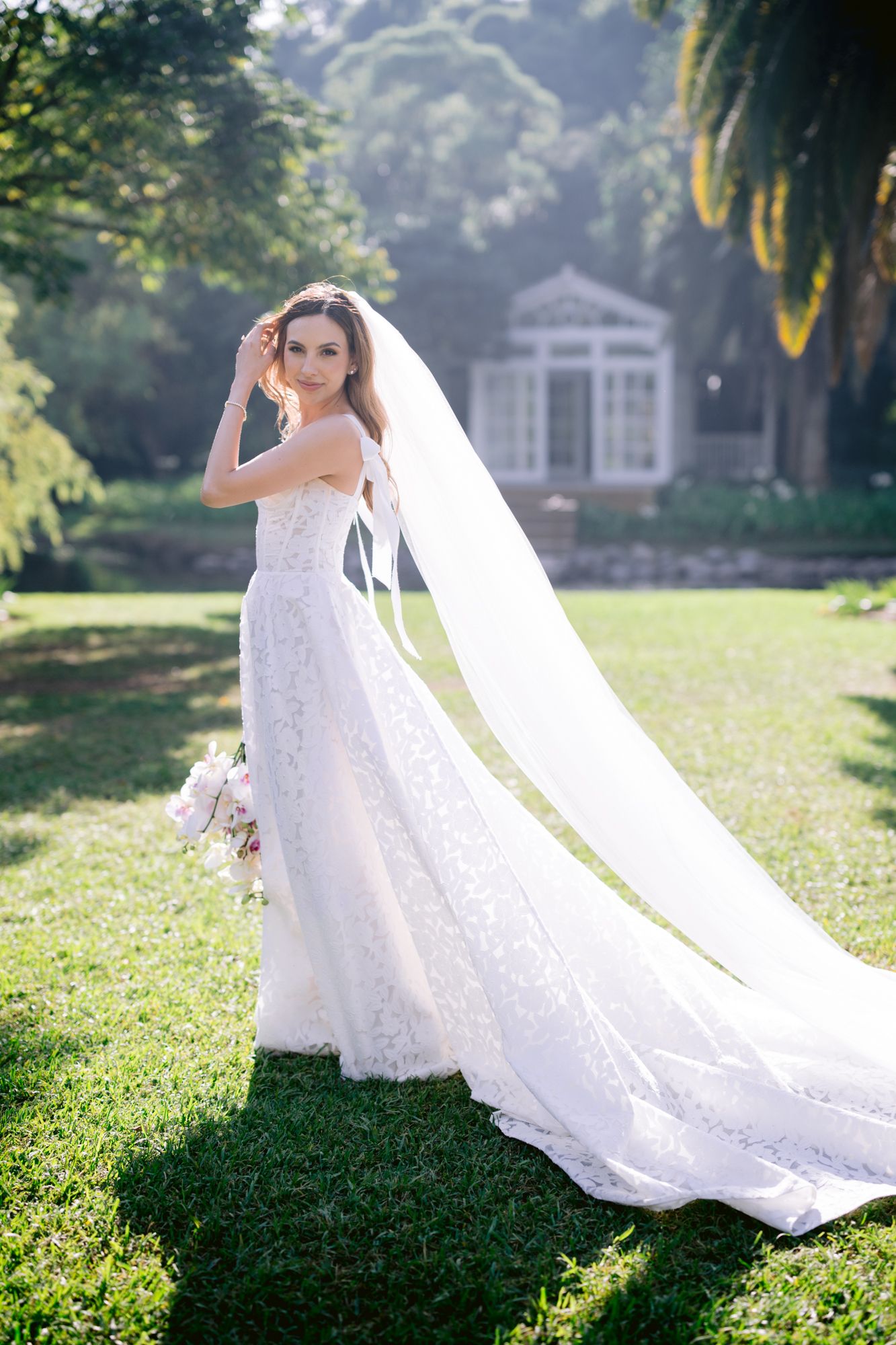 Bride in lace wedding gown with long flowing veil poses in sunlit garden with classic architecture.