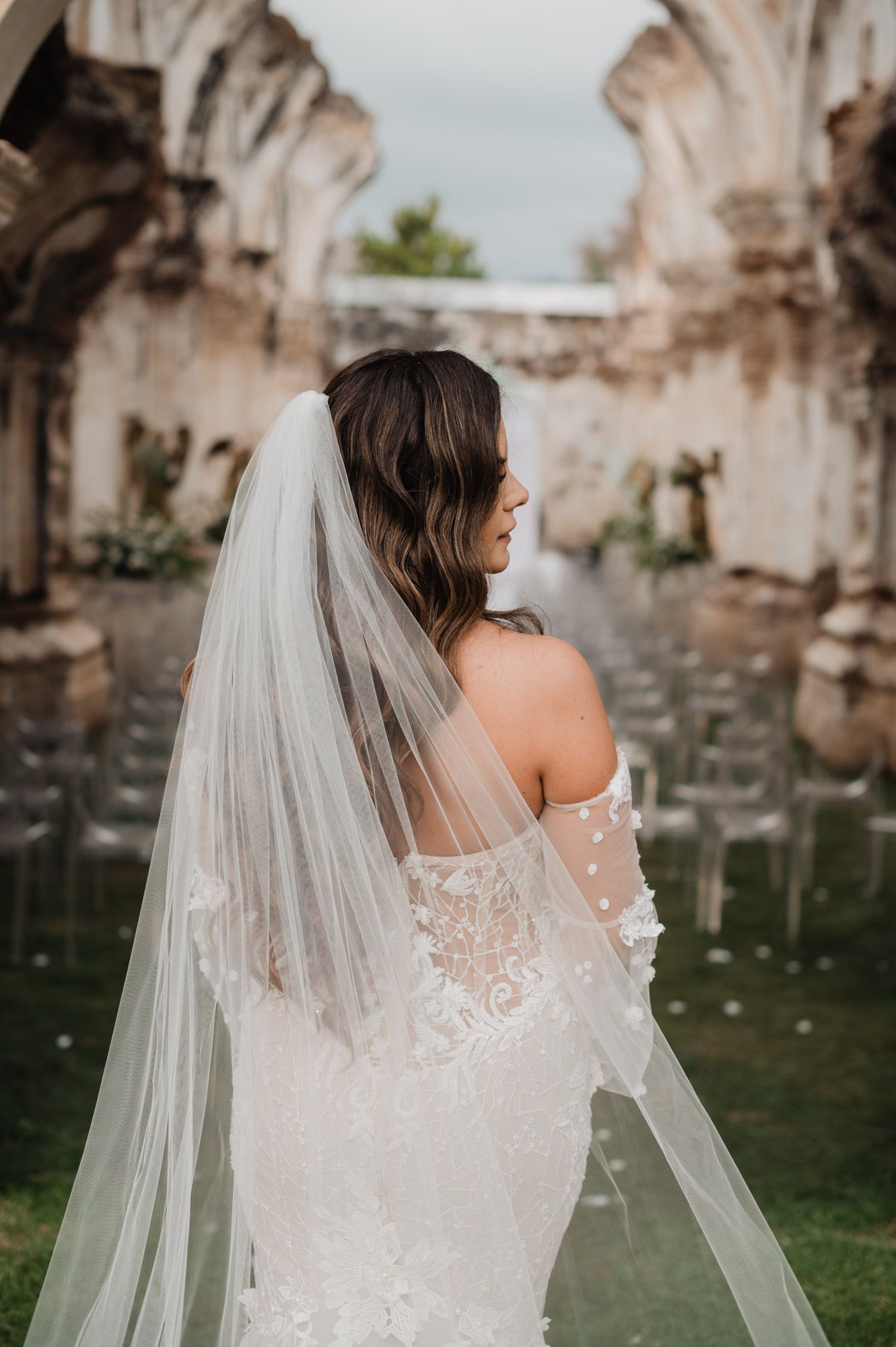 Delicate lace wedding dress and flowing veil detail against rustic stone ruins backdrop.
