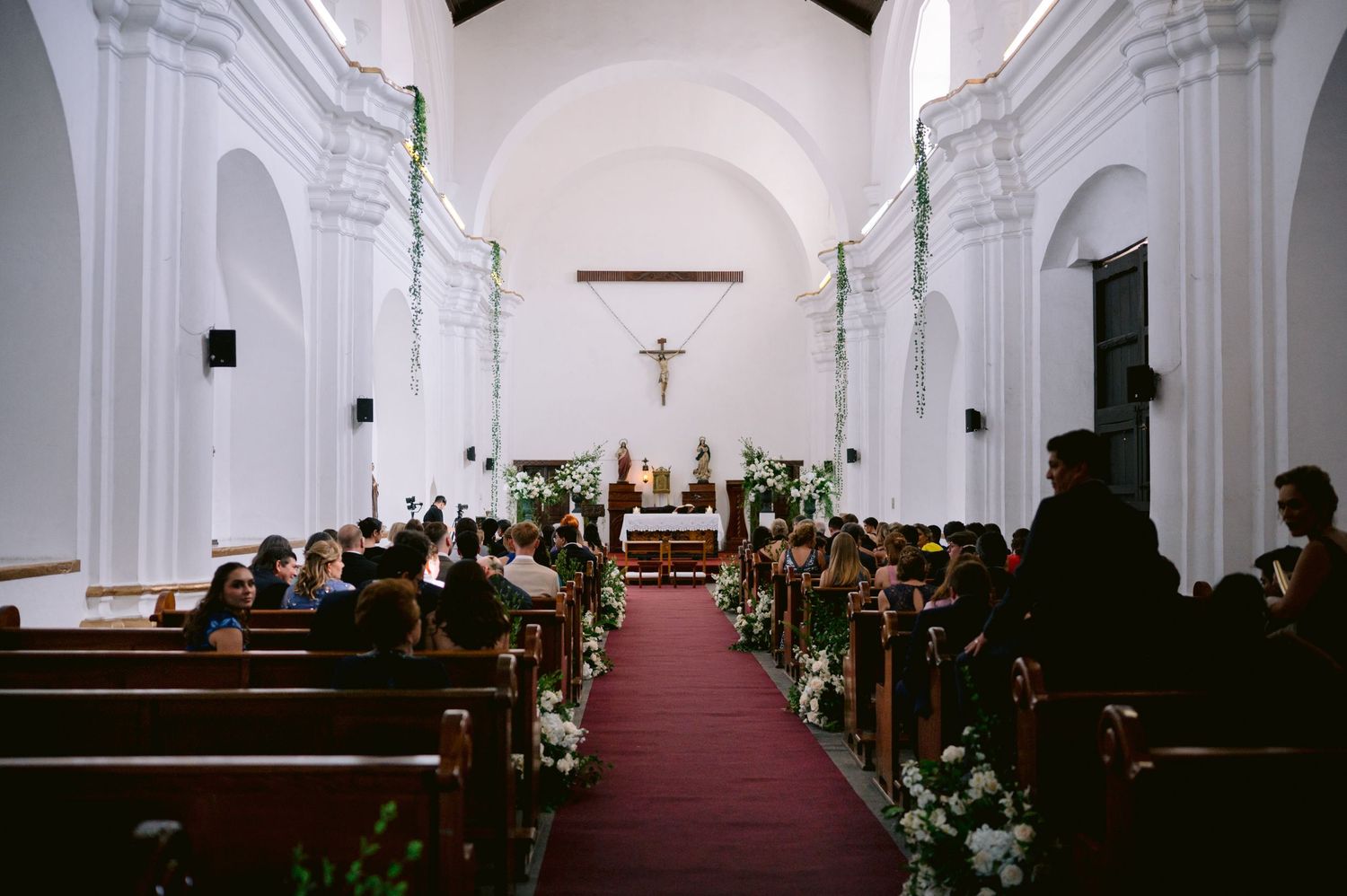 Church ceremony setup with floral decorations along a red-carpeted aisle in a white-walled sanctuary.