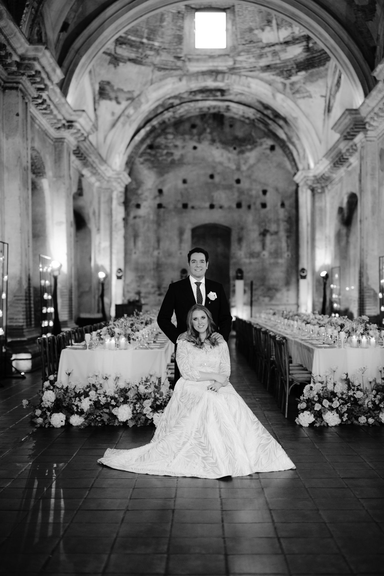 Wedding couple poses in historic church hall with decorated reception tables and floral arrangements.