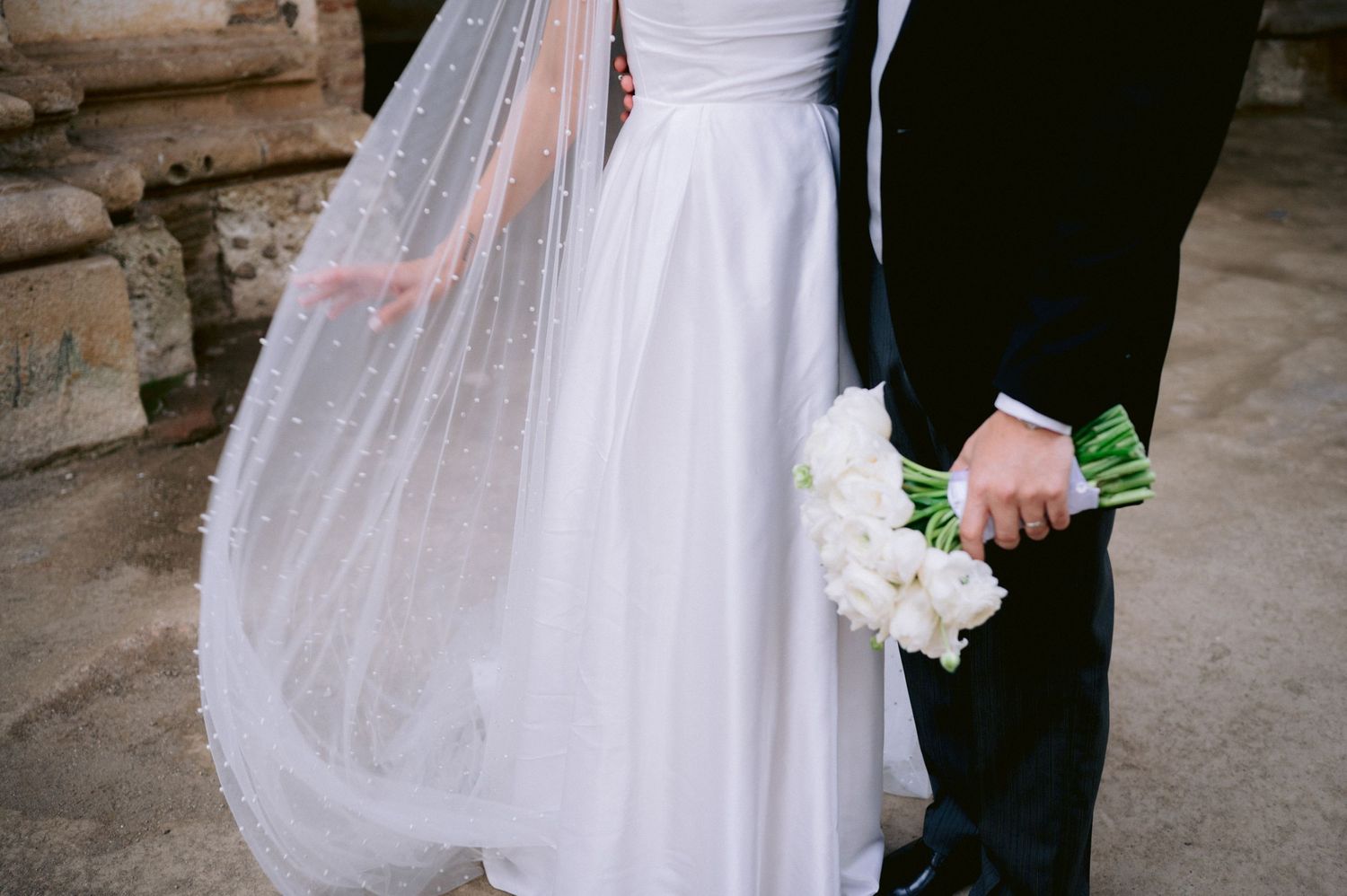 Detail shot of wedding couple with flowing veil and white bouquet against stone background.