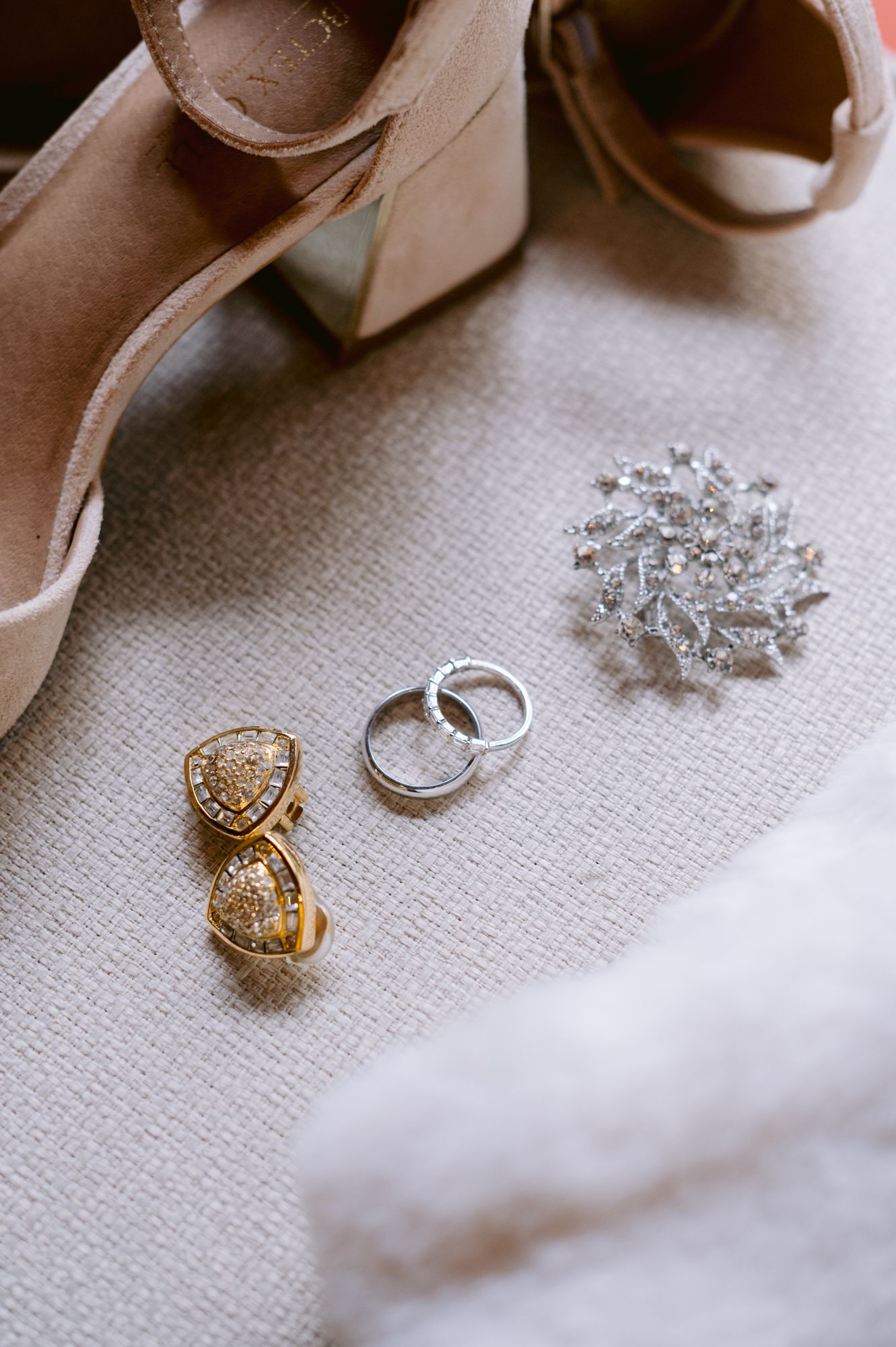 Wedding accessories including gold earrings, silver rings and a brooch arranged with beige shoes.