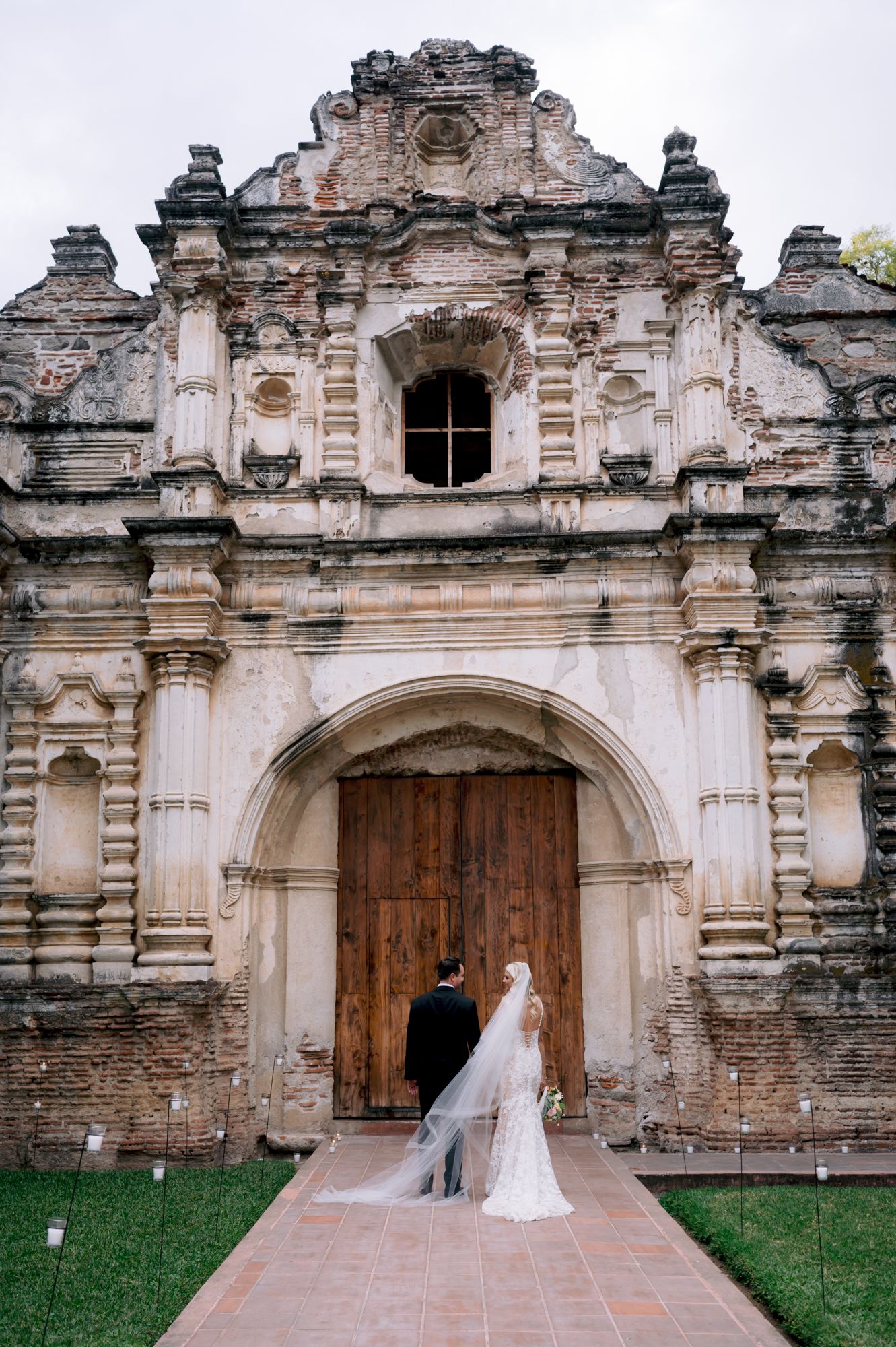 A wedding couple framed by the grand entrance of a Spanish colonial church with stone architecture.