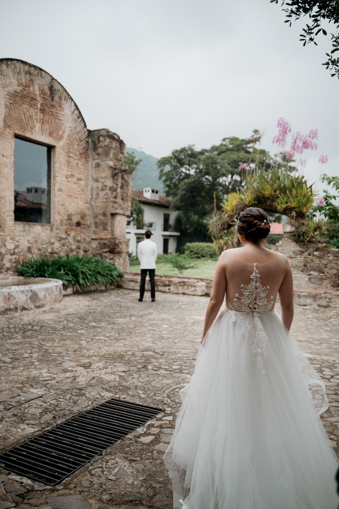 Bride in lace-back wedding gown approaches figure in white suit for first look moment.