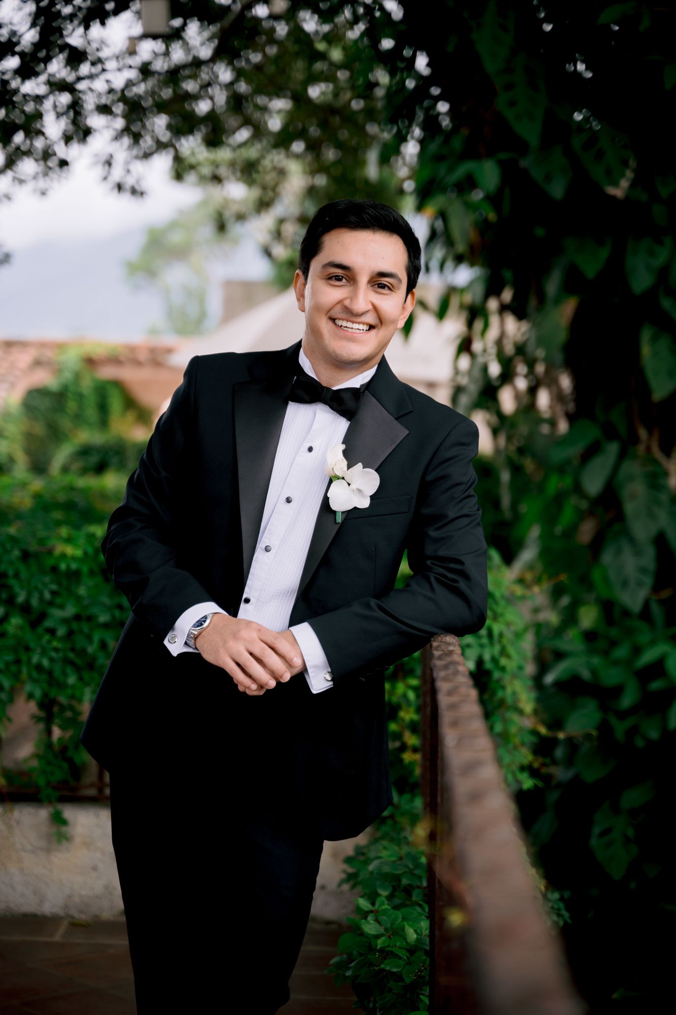 Groom in classic black tuxedo and bow tie poses in front of garden greenery.