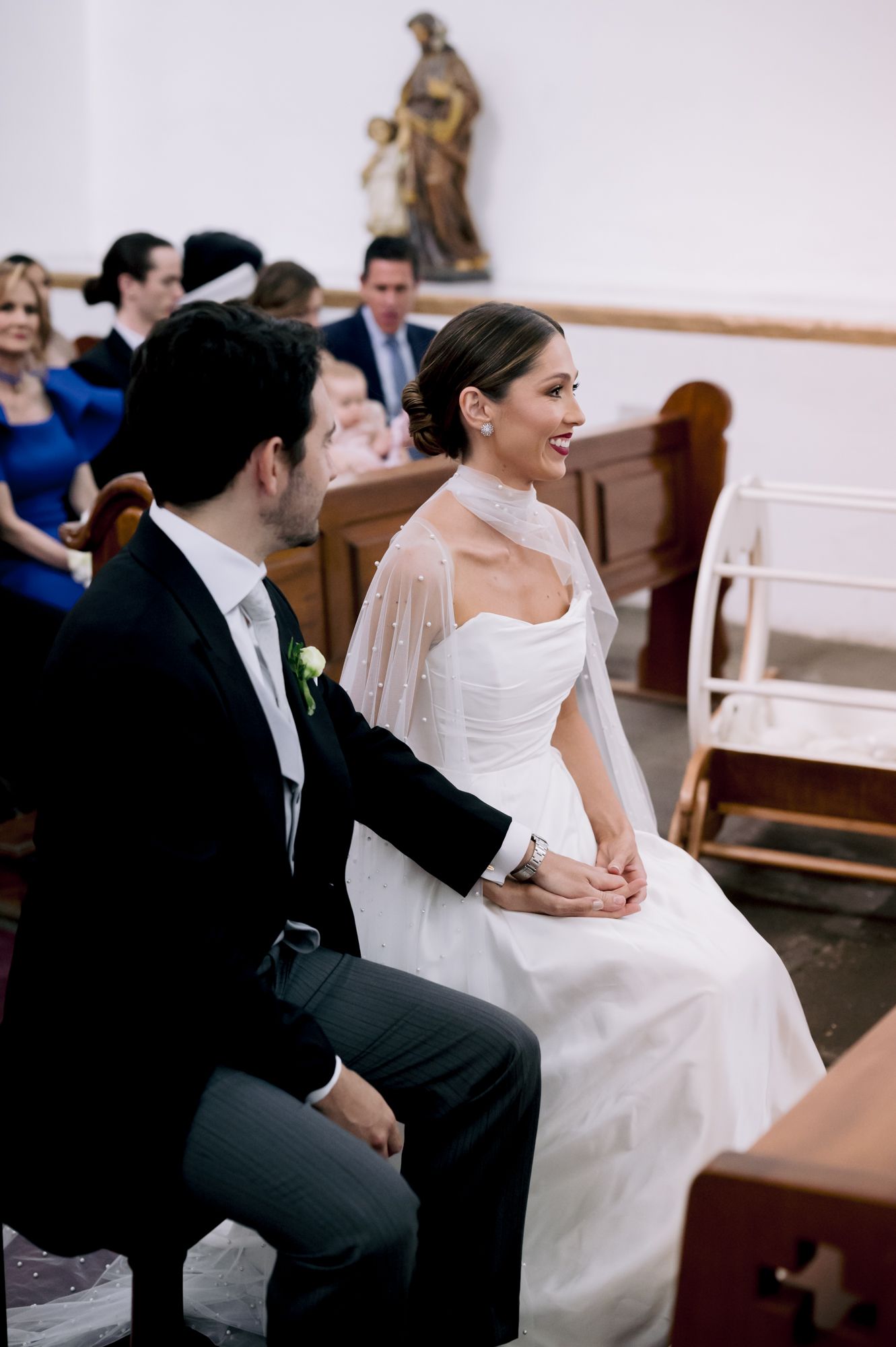 Newlyweds seated in church pews during wedding ceremony with guests in background.