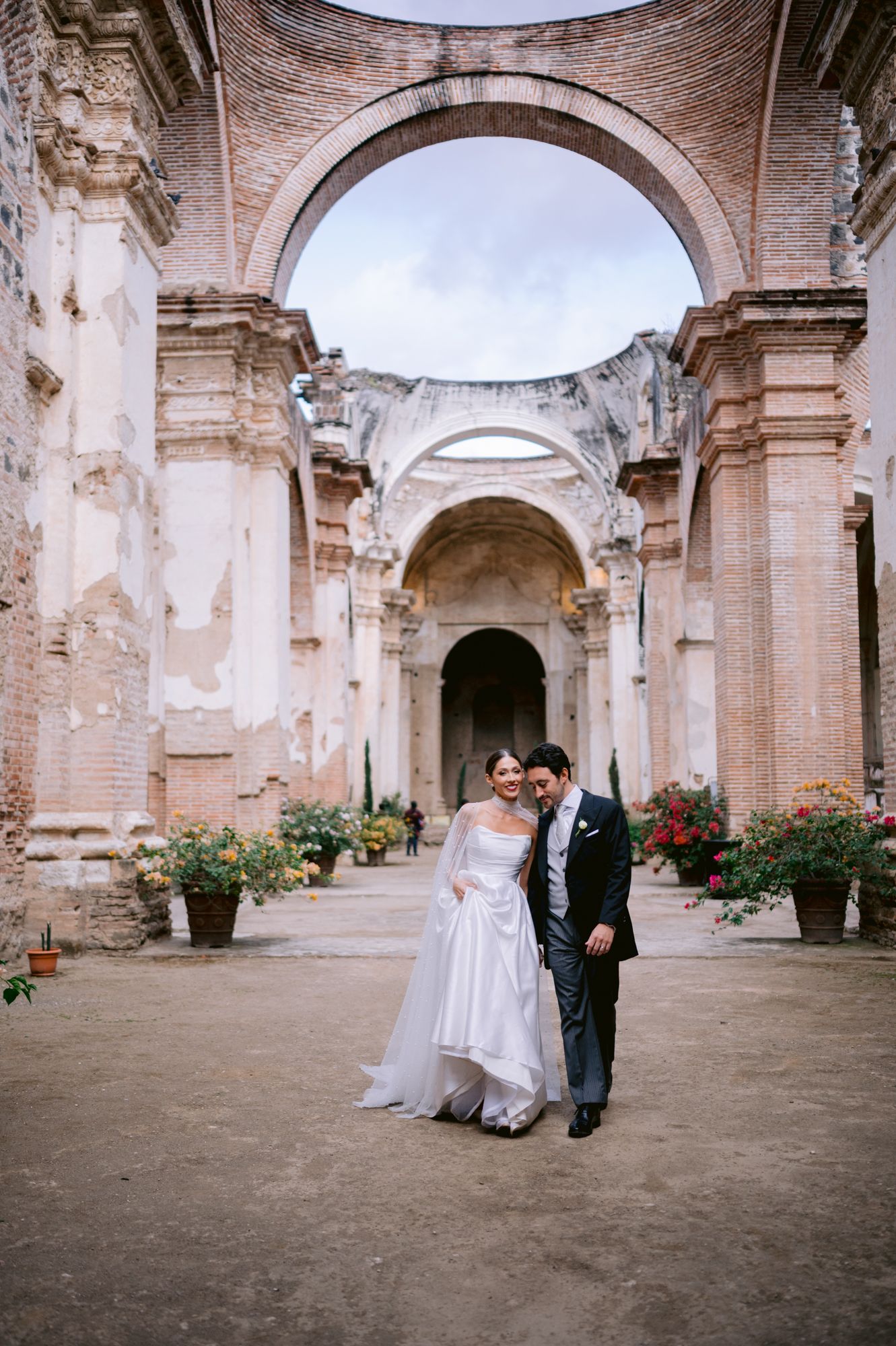 Wedding couple walks together through grand arched ruins with potted flowers.
