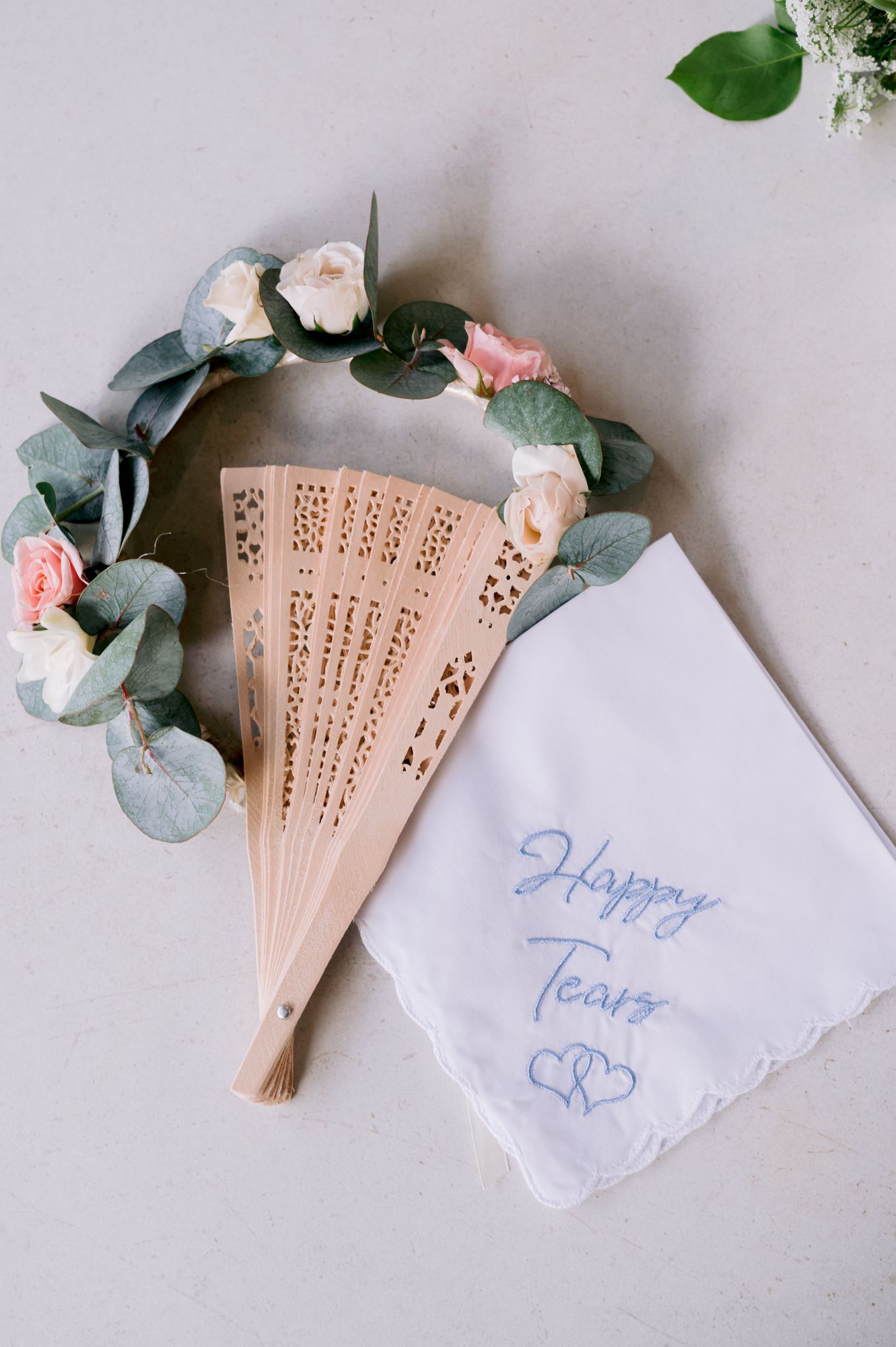 Delicate wedding accessories including a floral wreath and decorative fan with 'Happy Tears' handkerchief.