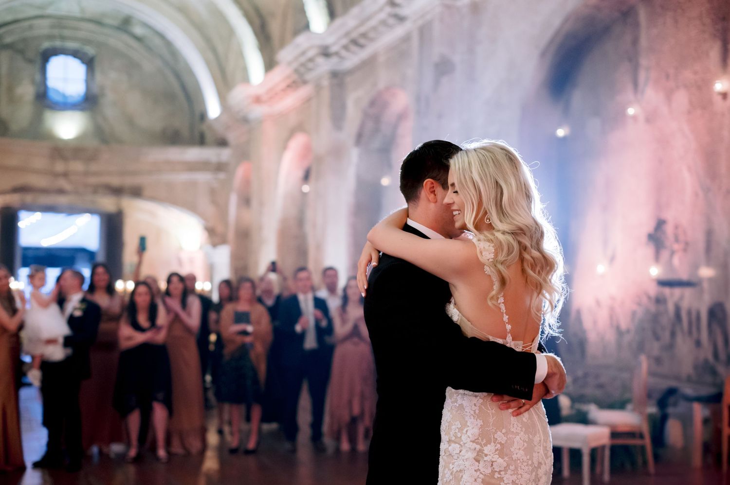 Dancing couple celebrate their first dance in an elegant stone venue with guests watching.