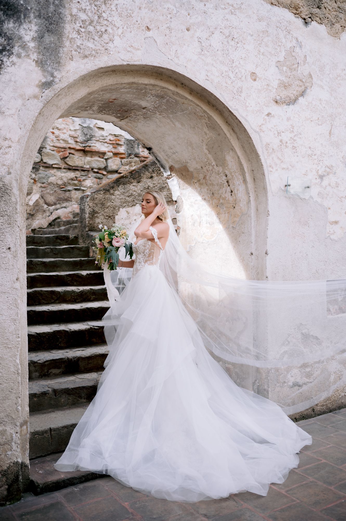 Ethereal bridal portrait with flowing veil catching wind under historic stone archway.