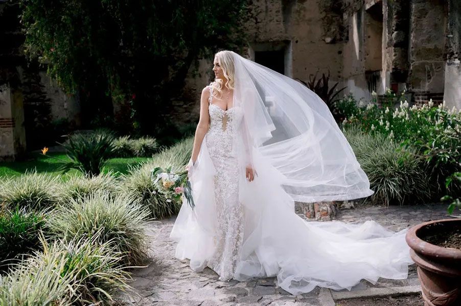 A bride in a flowing lace gown and dramatic long veil stands in a garden courtyard with potted plants and foliage.