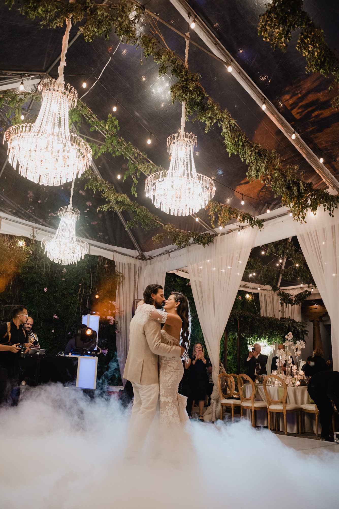 Newlyweds share first dance under crystal chandeliers with dramatic fog effect on dance floor.