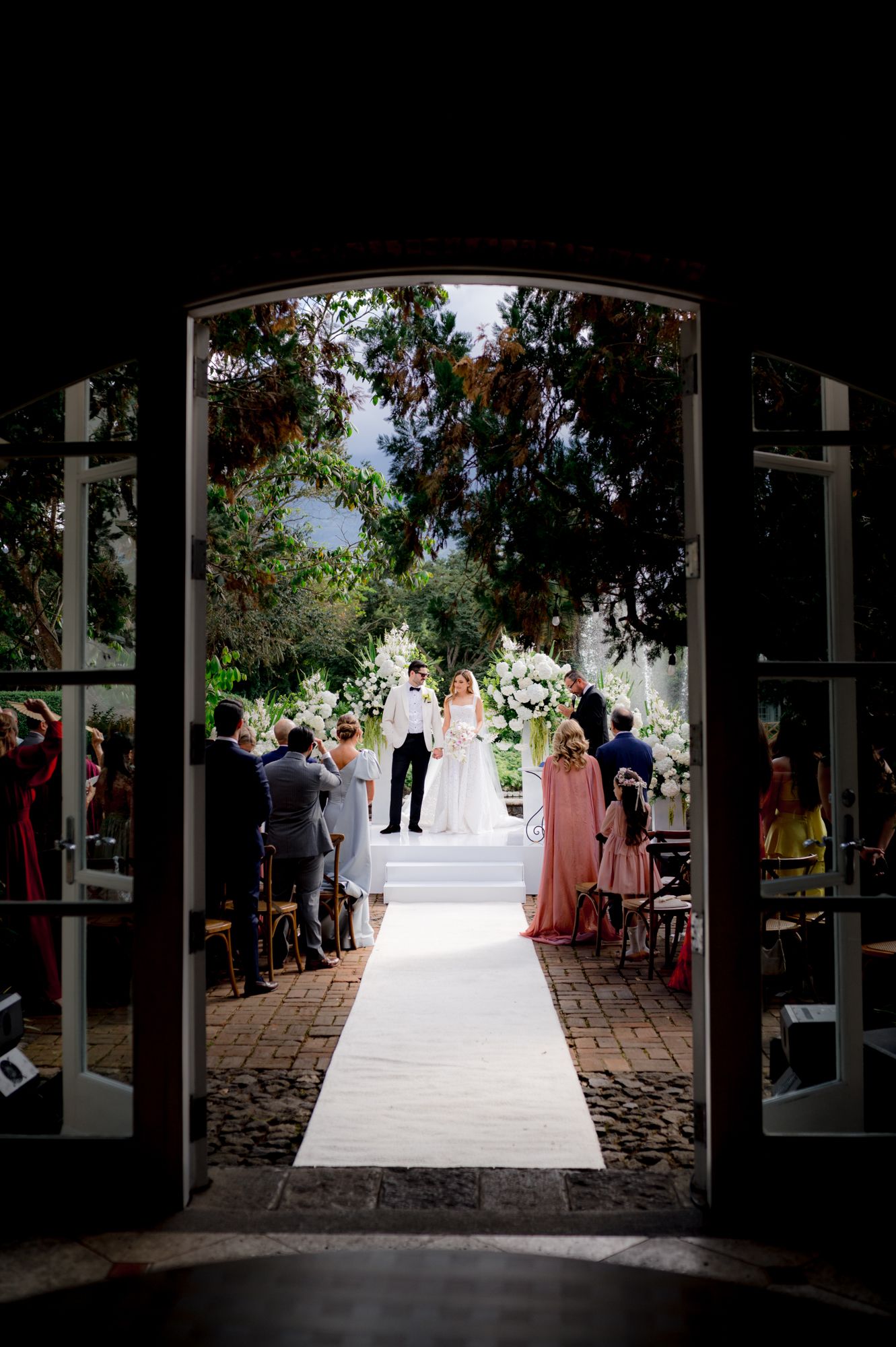 Wedding ceremony setup viewed through elegant French doors with a white aisle runner and guests seated.