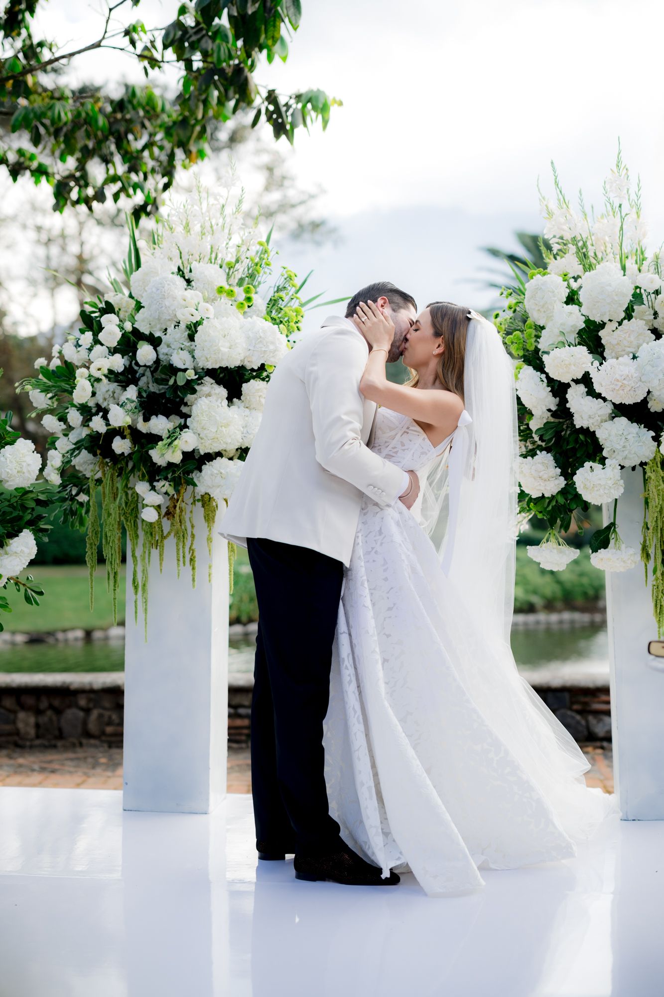 Newlyweds share their first kiss at outdoor wedding ceremony decorated with white flowers and columns.