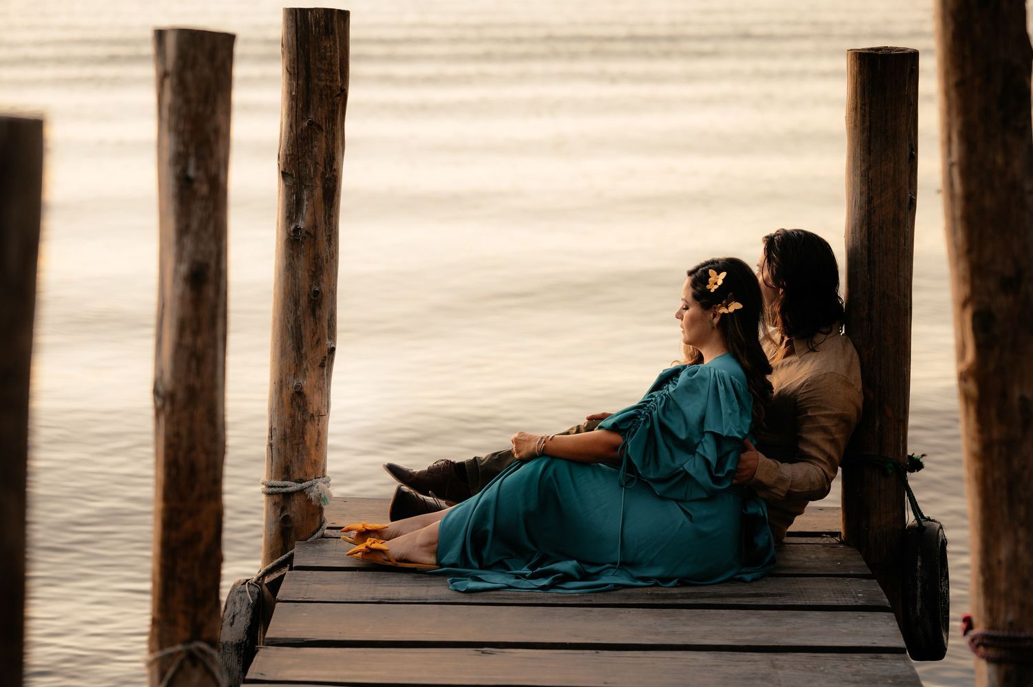 People relax on a wooden pier extending over calm water during sunset, creating long shadows on the deck.