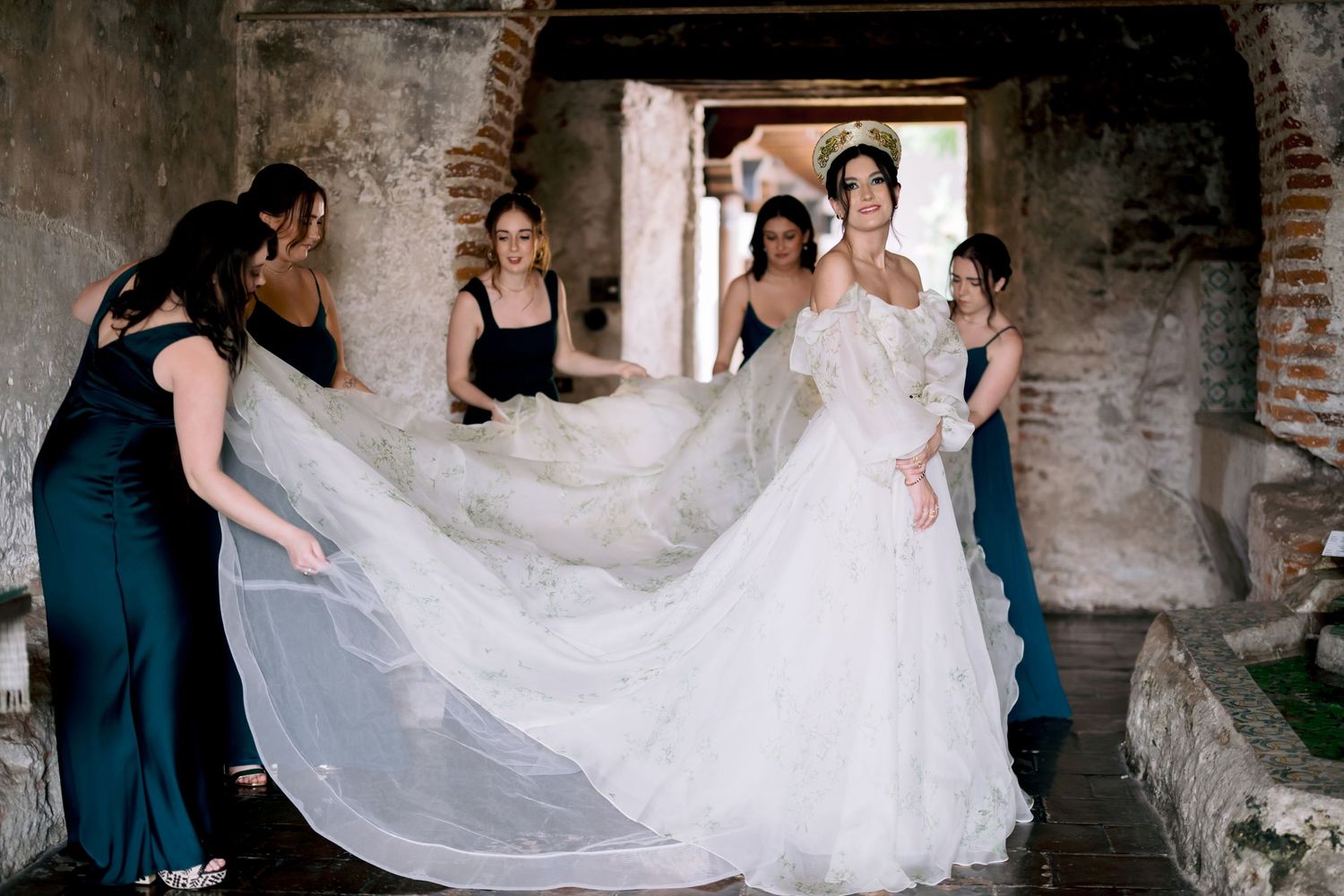 Bridesmaids in blue dresses help arrange a flowing wedding dress train in a rustic setting.