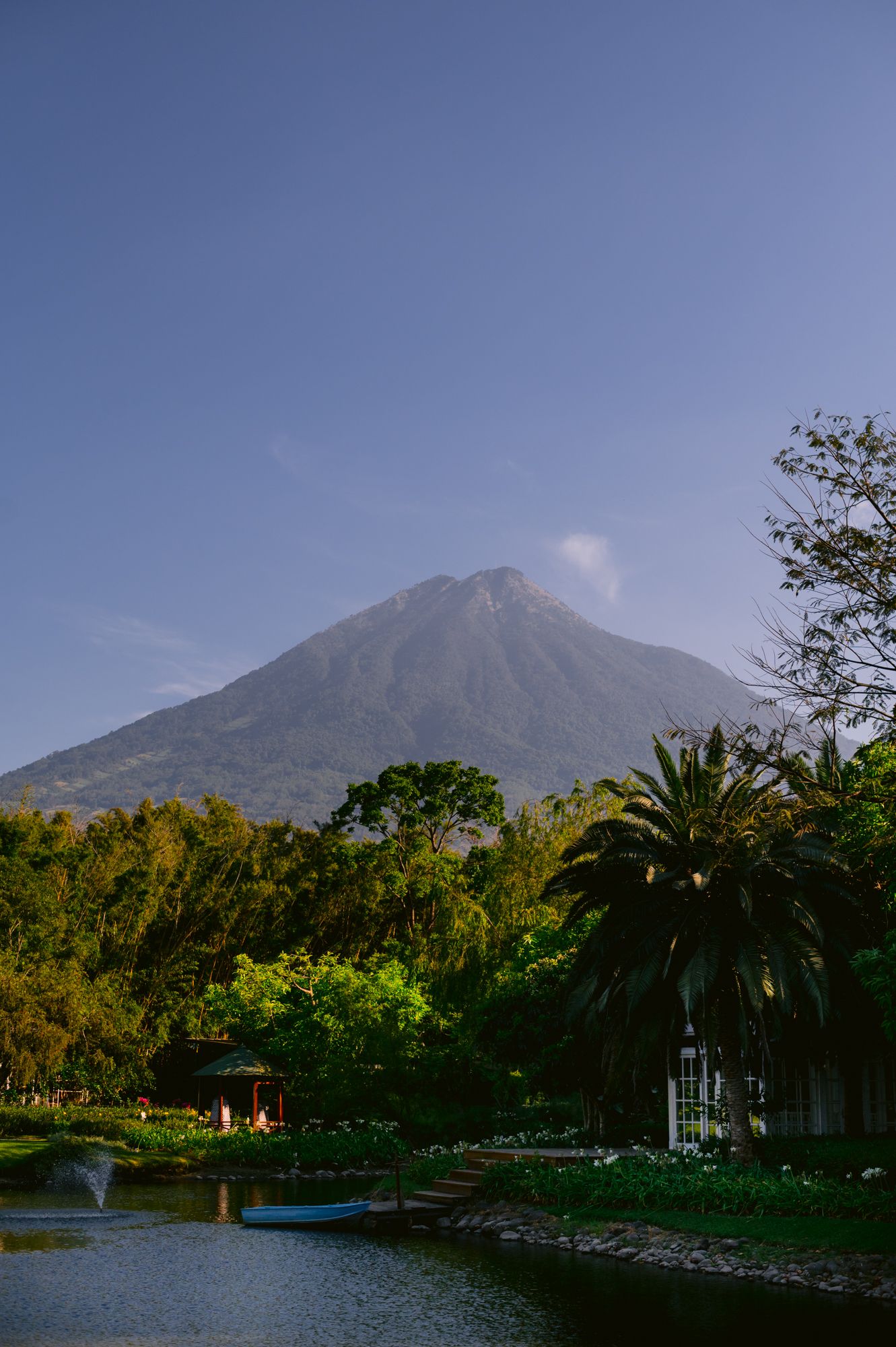 A majestic volcanic mountain rises above a lush landscape with a tranquil pond and palm trees in the foreground.