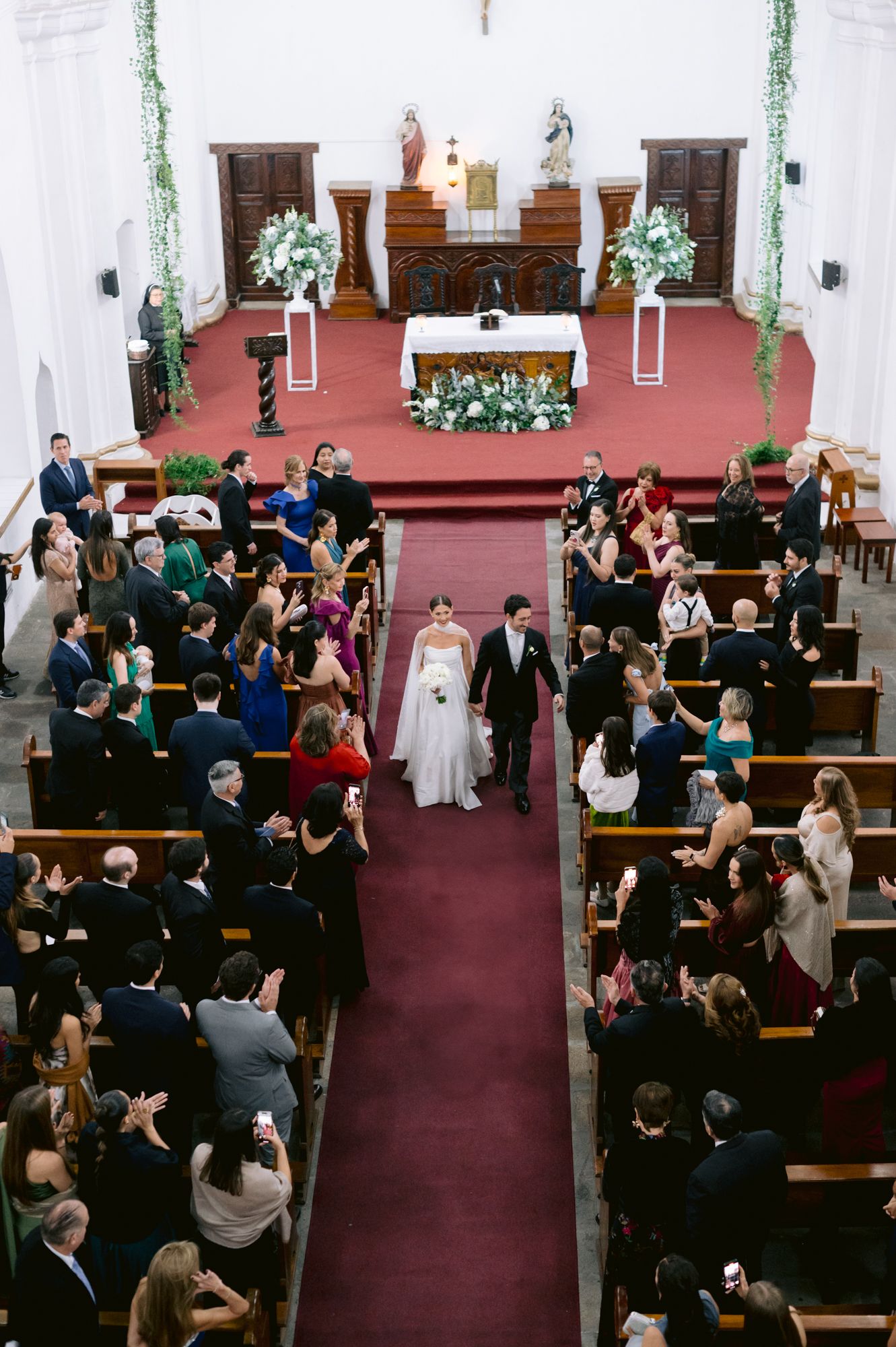 Aerial view of wedding ceremony in traditional church filled with guests and decorated with flowers.