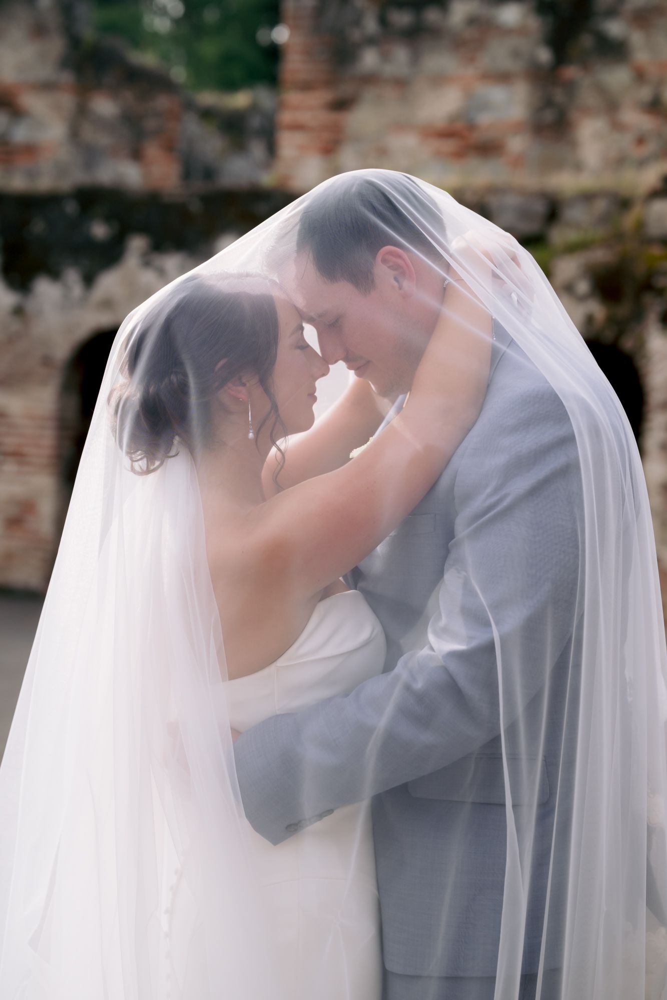 Romantic moment captured under flowing bridal veil against rustic stone architecture.