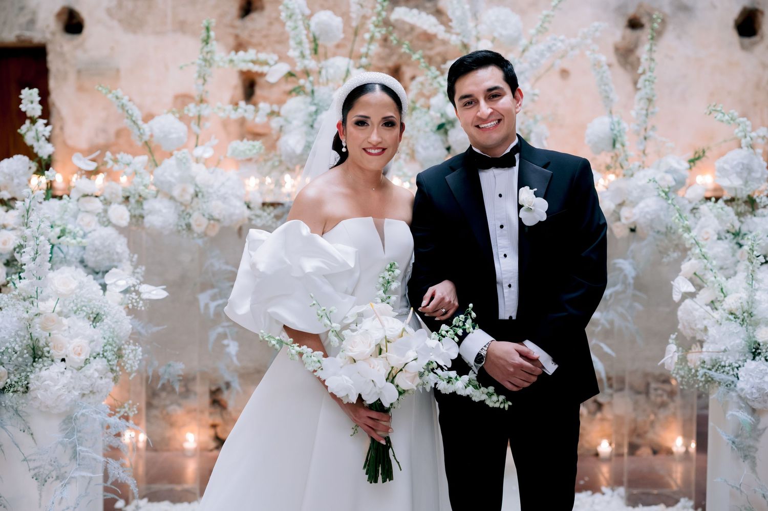 Wedding couple stands among elegant white floral arrangements and soft lighting at luxury reception venue.