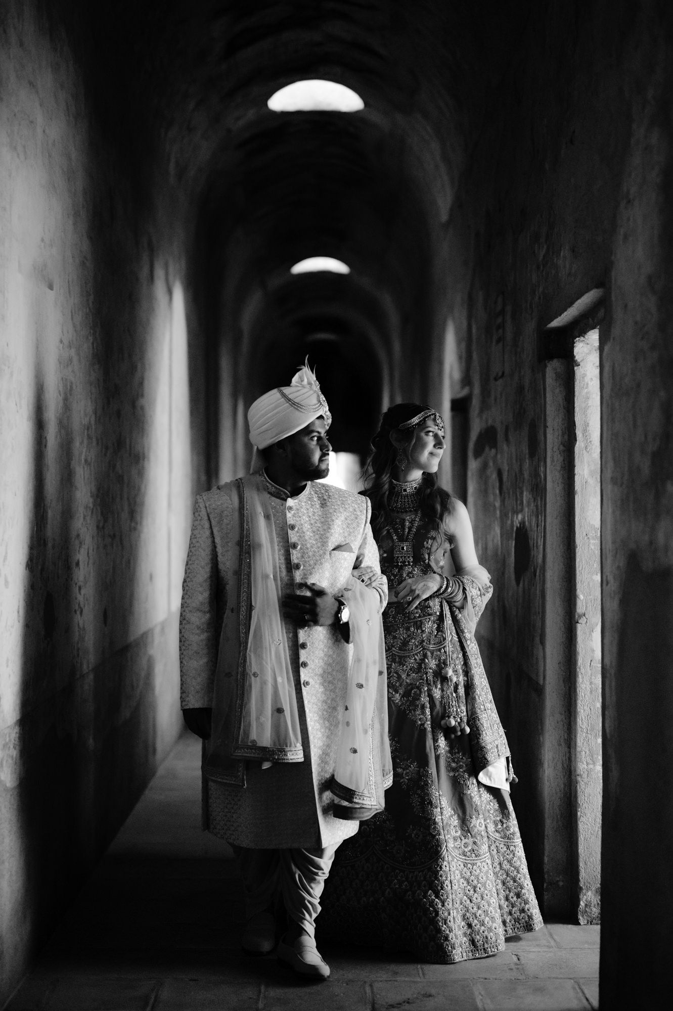Black and white photo of couple in traditional wedding attire standing in dramatic corridor.