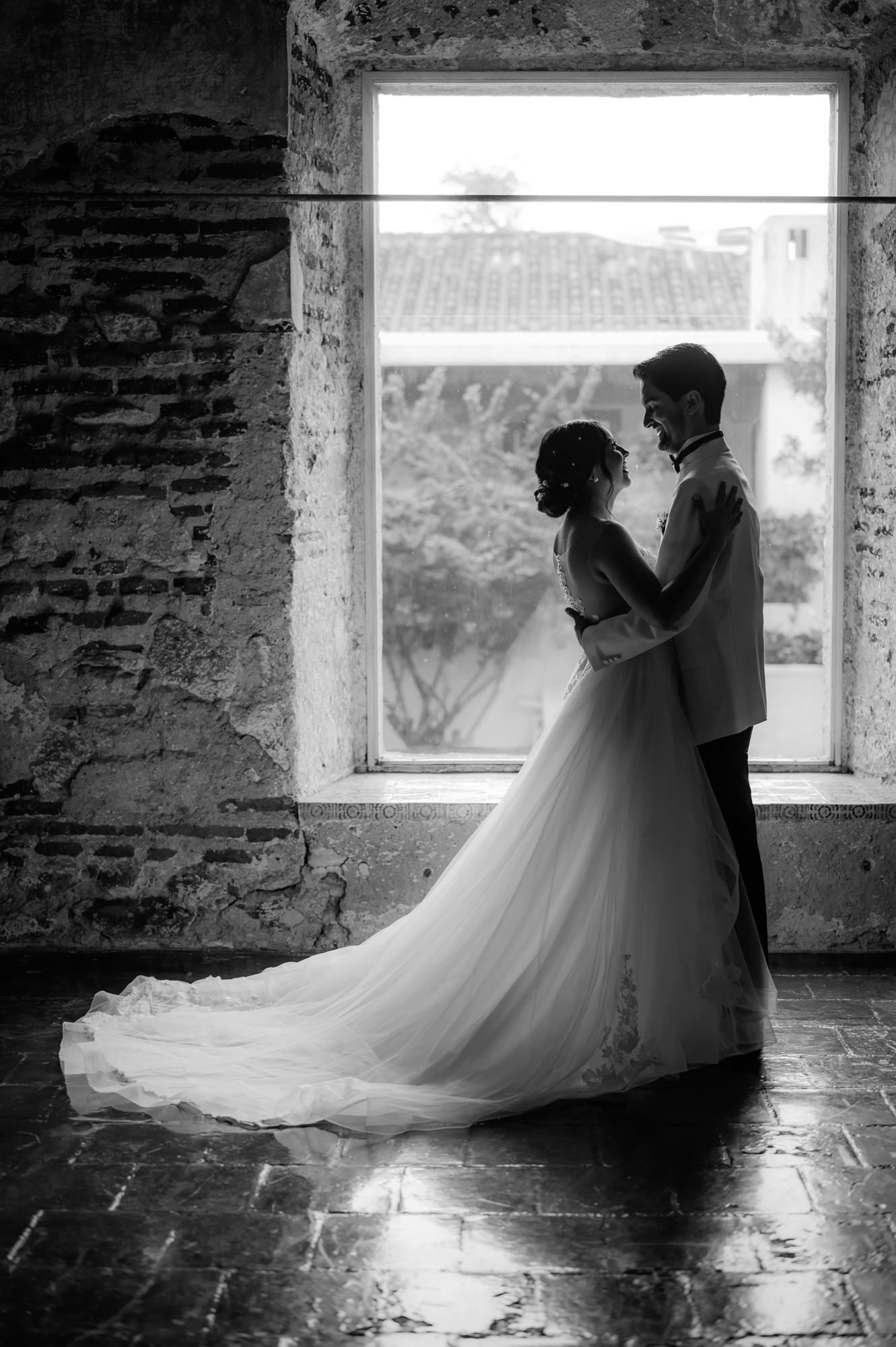 A romantic black and white silhouette of a couple in wedding attire standing by a rustic window.
