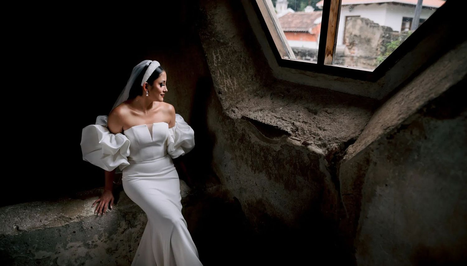 Elegant white wedding dress photographed in a rustic stone window alcove with rooftops visible in background.