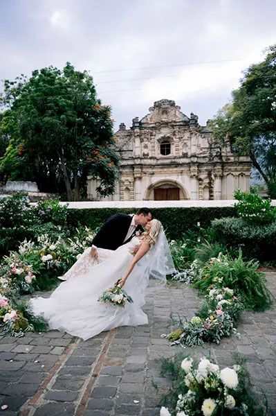 A romantic wedding moment in front of an ornate colonial church facade with floral decorations on cobblestone ground.