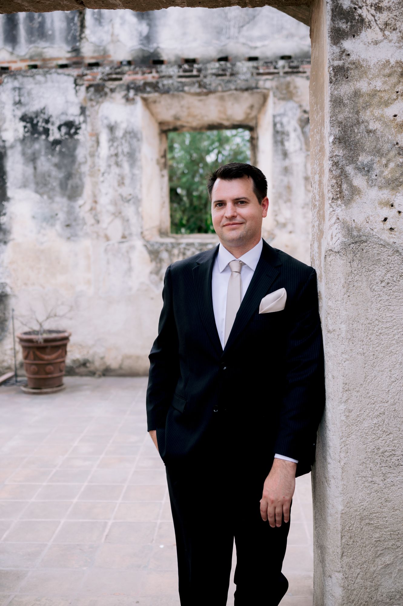 Groom in classic black tuxedo poses against weathered stone wall in historic setting.