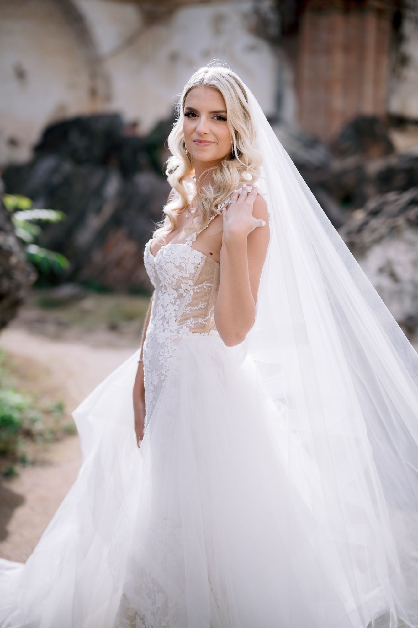 A bride in an elegant lace wedding dress with flowing veil poses against ancient stone ruins.
