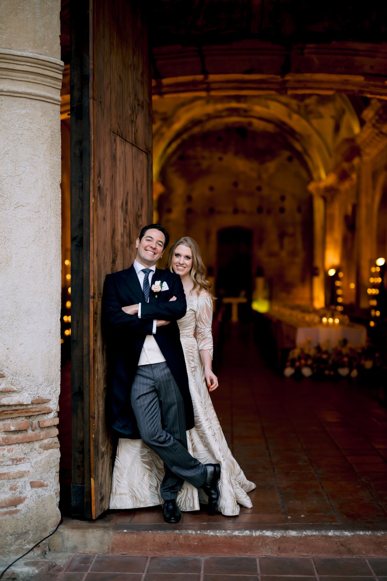 Wedding couple poses playfully against a historic stone column in front of ornate archways.