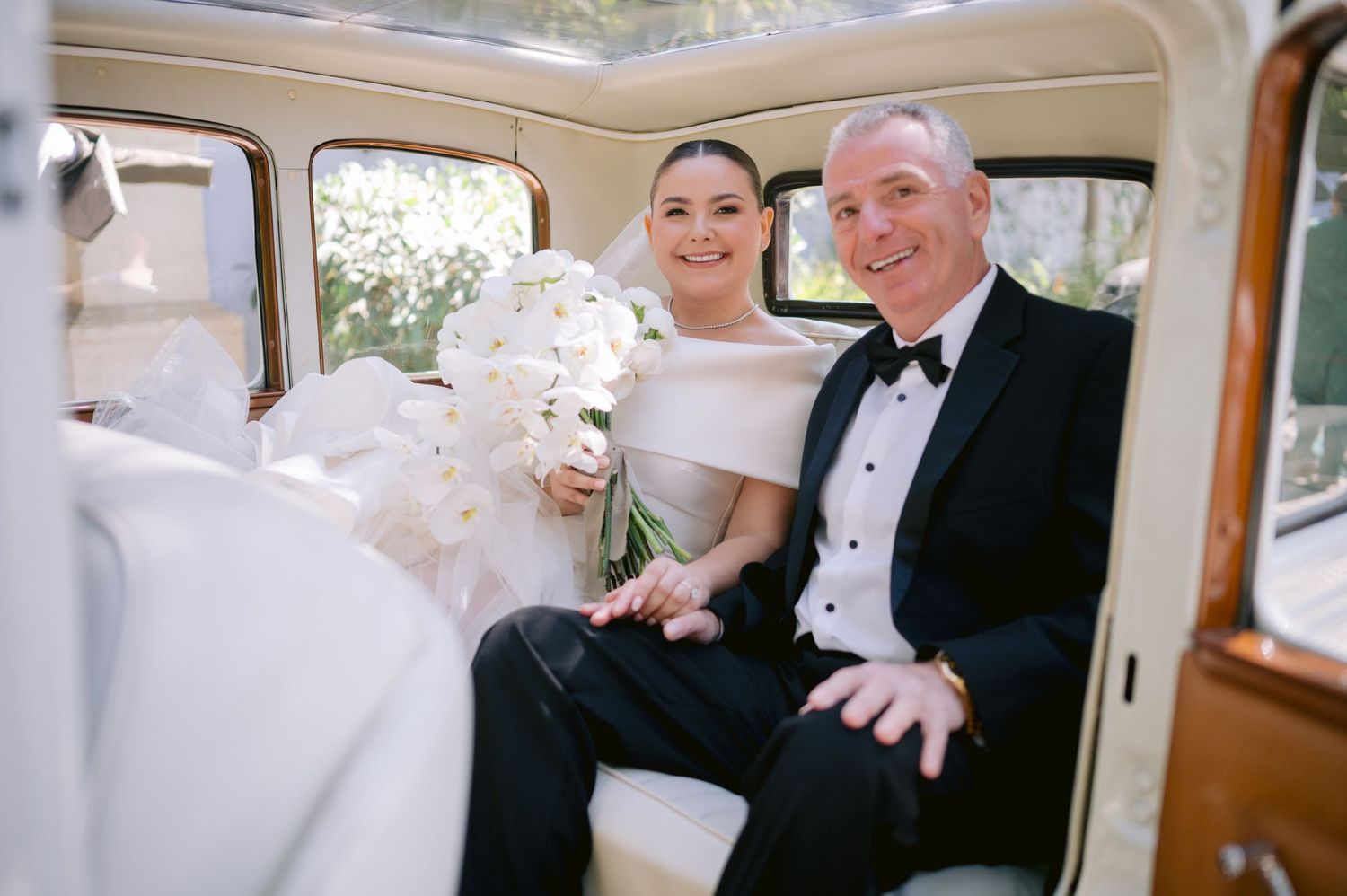 Happy newlyweds sitting inside a classic vintage car with white bouquet.