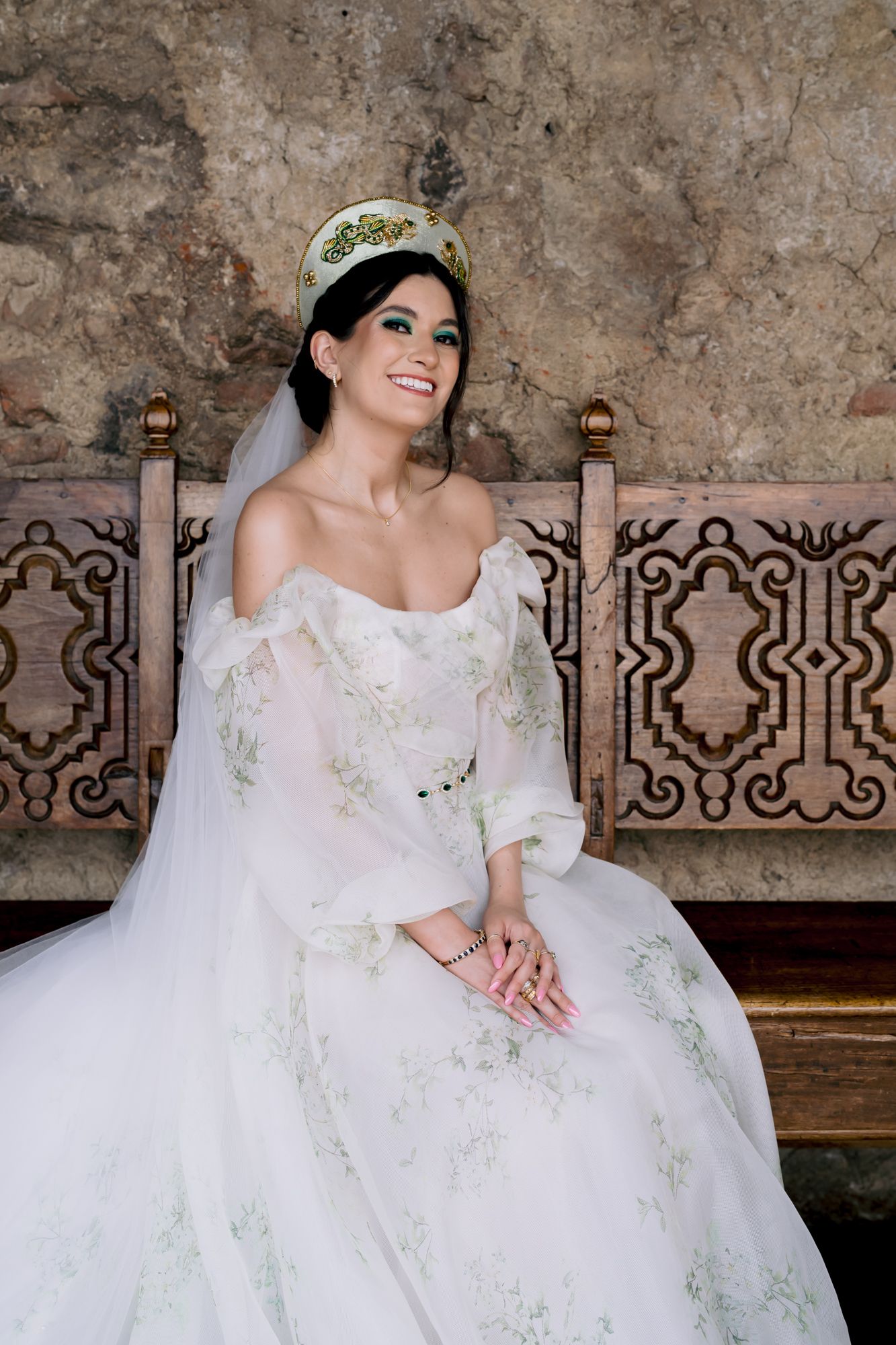 Elegant wedding gown with delicate floral embroidery and off-shoulder design against rustic stone wall backdrop.