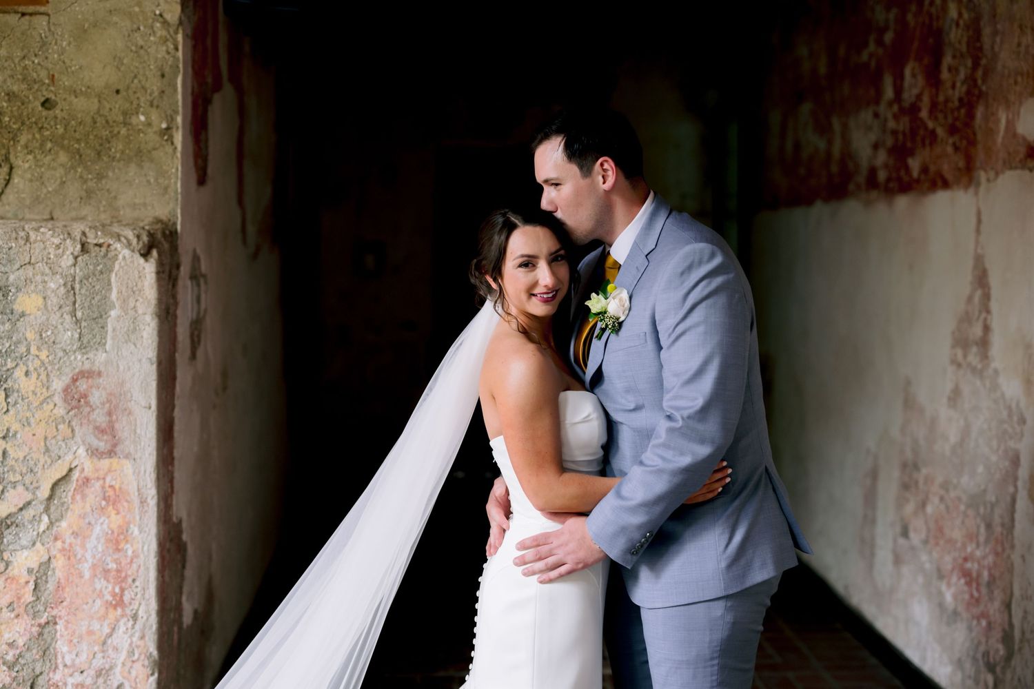 Wedding portrait of a couple embracing in front of a weathered stone wall with romantic lighting.