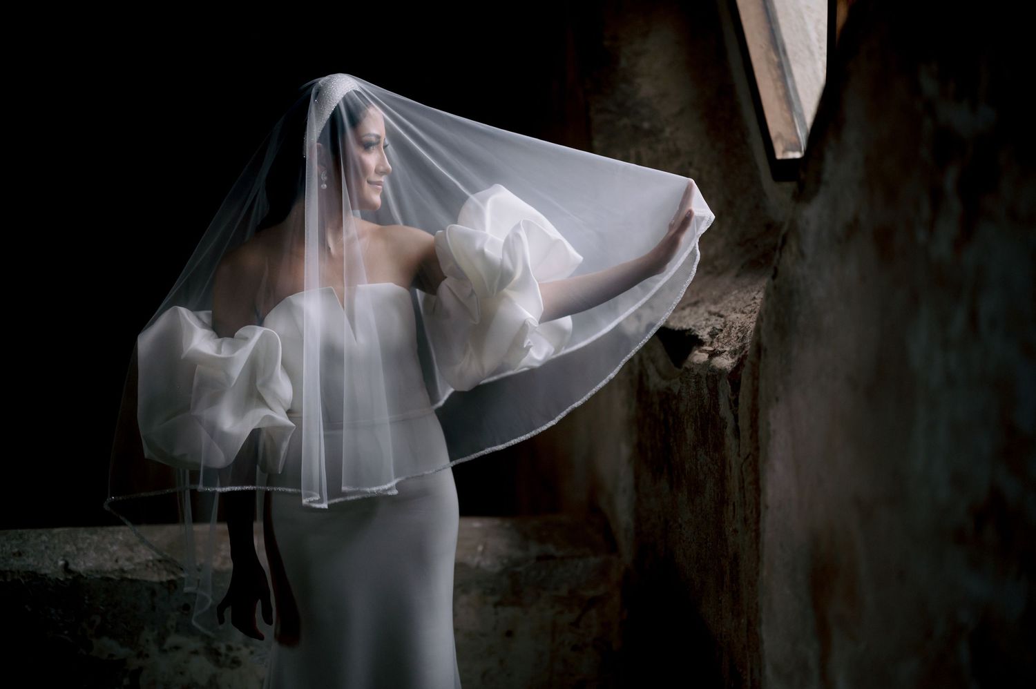 Artistic bridal portrait with flowing veil creating movement in shadowy architectural setting.
