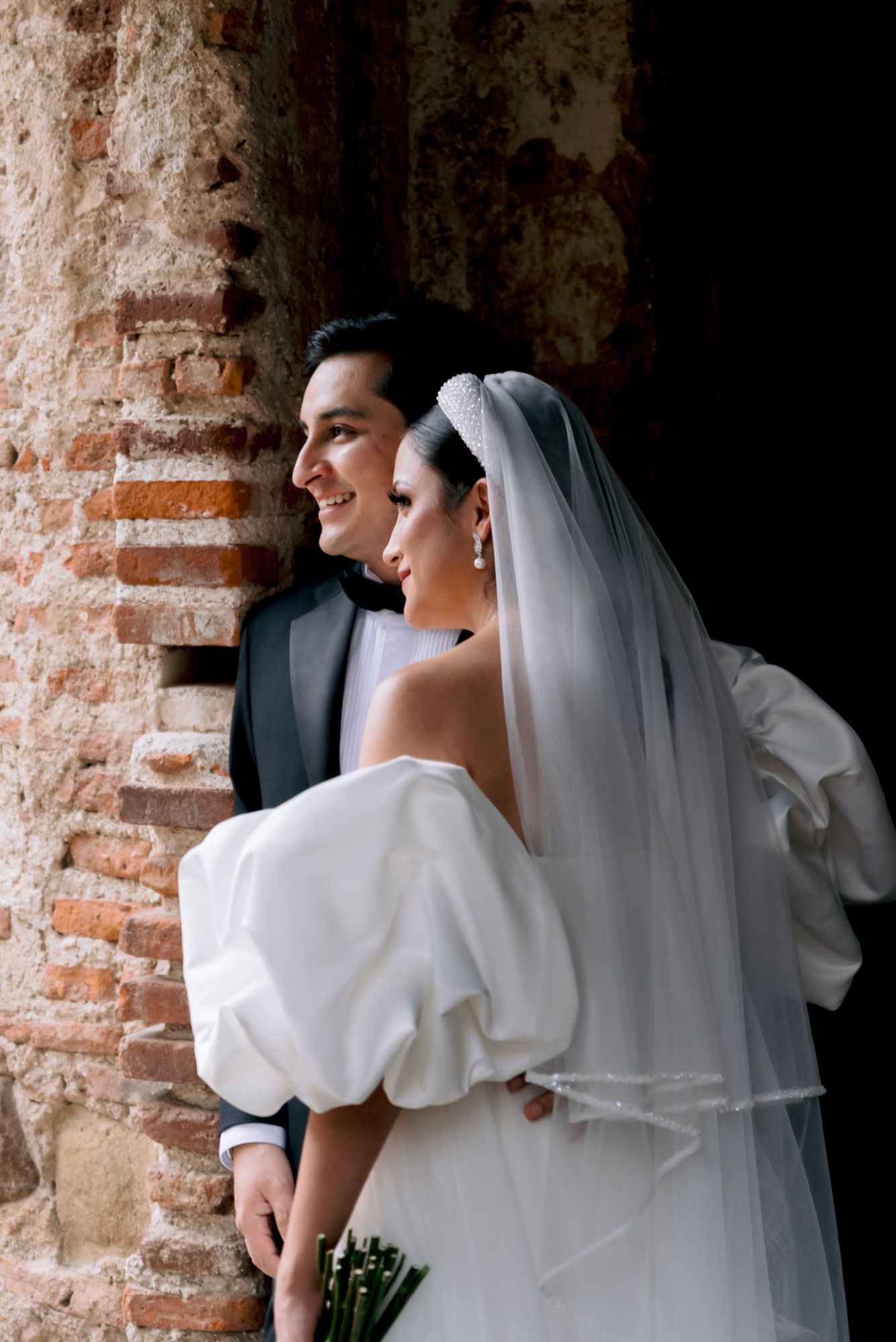 Elegant wedding couple pose against rustic brick wall with bride wearing puffy-sleeved gown and flowing veil.