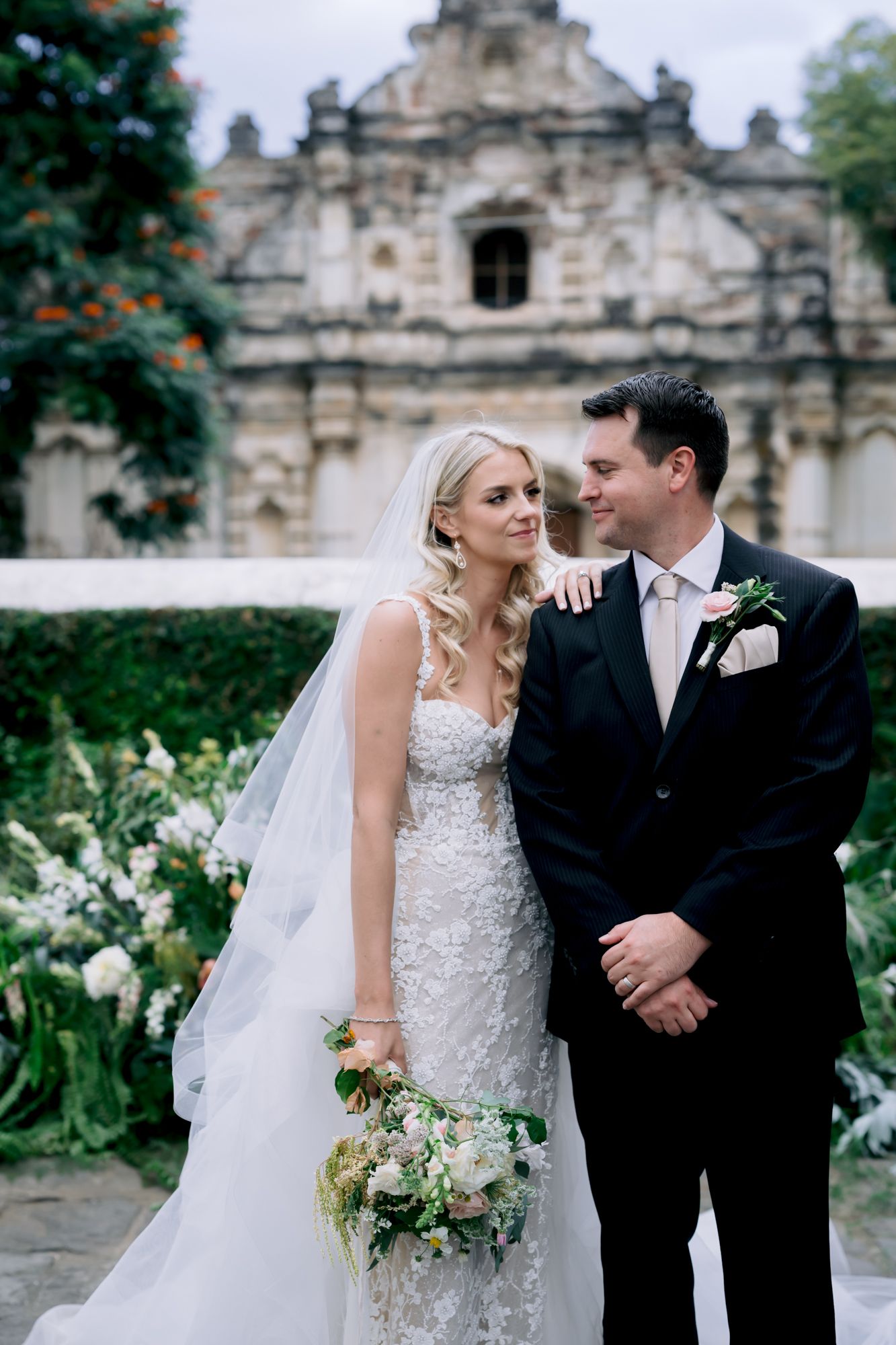 A romantic wedding portrait in front of a historic stone church facade with flowering gardens.