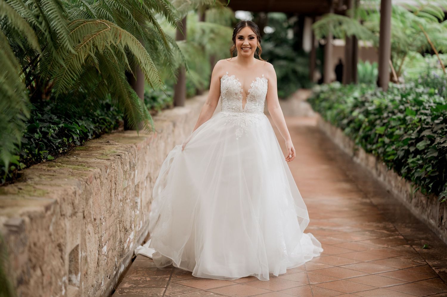 A bride in a flowing white ball gown poses on a stone-lined garden path surrounded by lush greenery.
