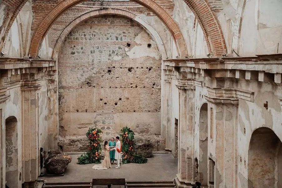 Couple posing under ancient stone arches of a weathered church ruins with ornate architectural details.