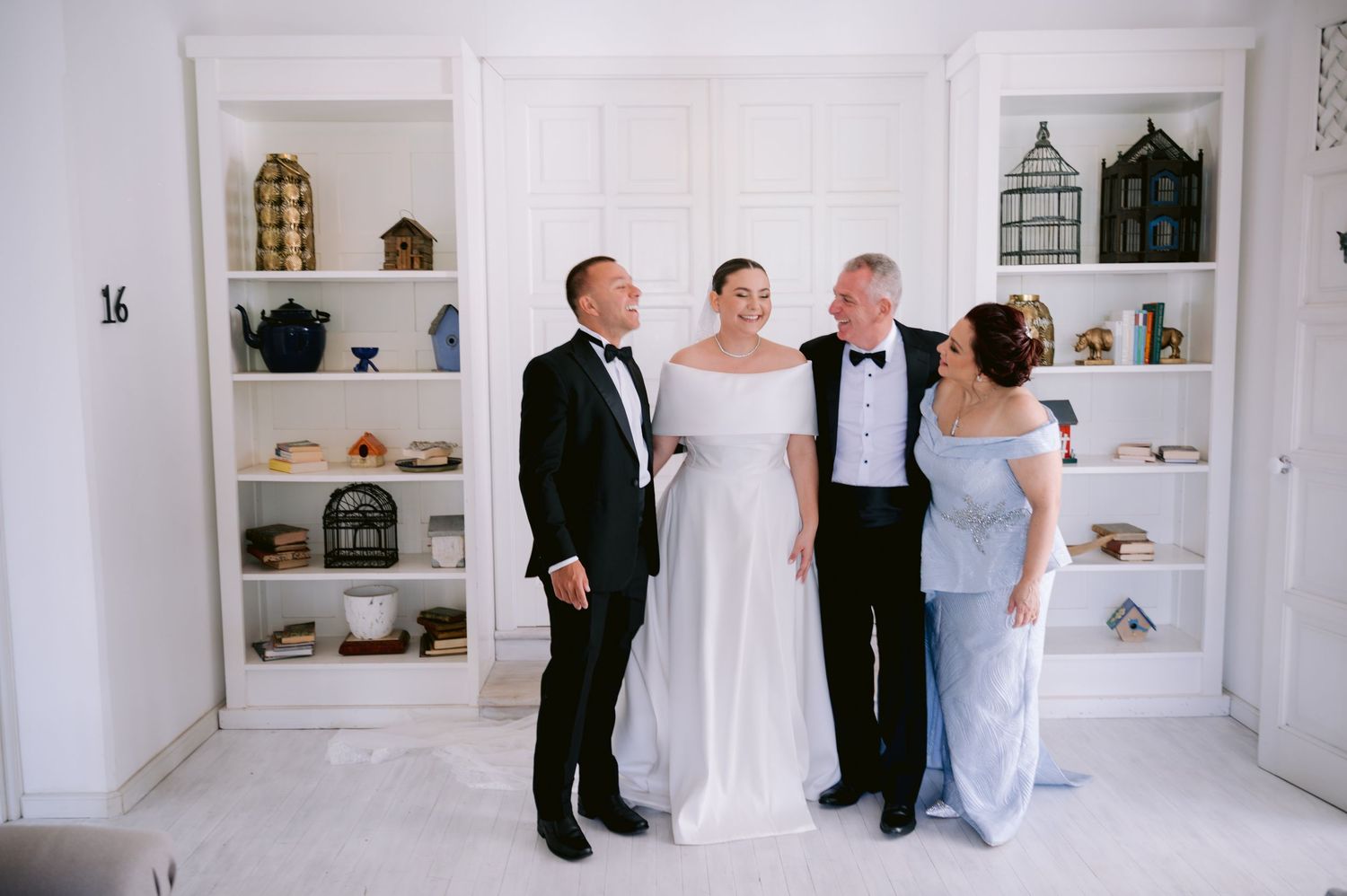 Wedding party poses in formal attire against white built-in shelves decorated with vintage items.