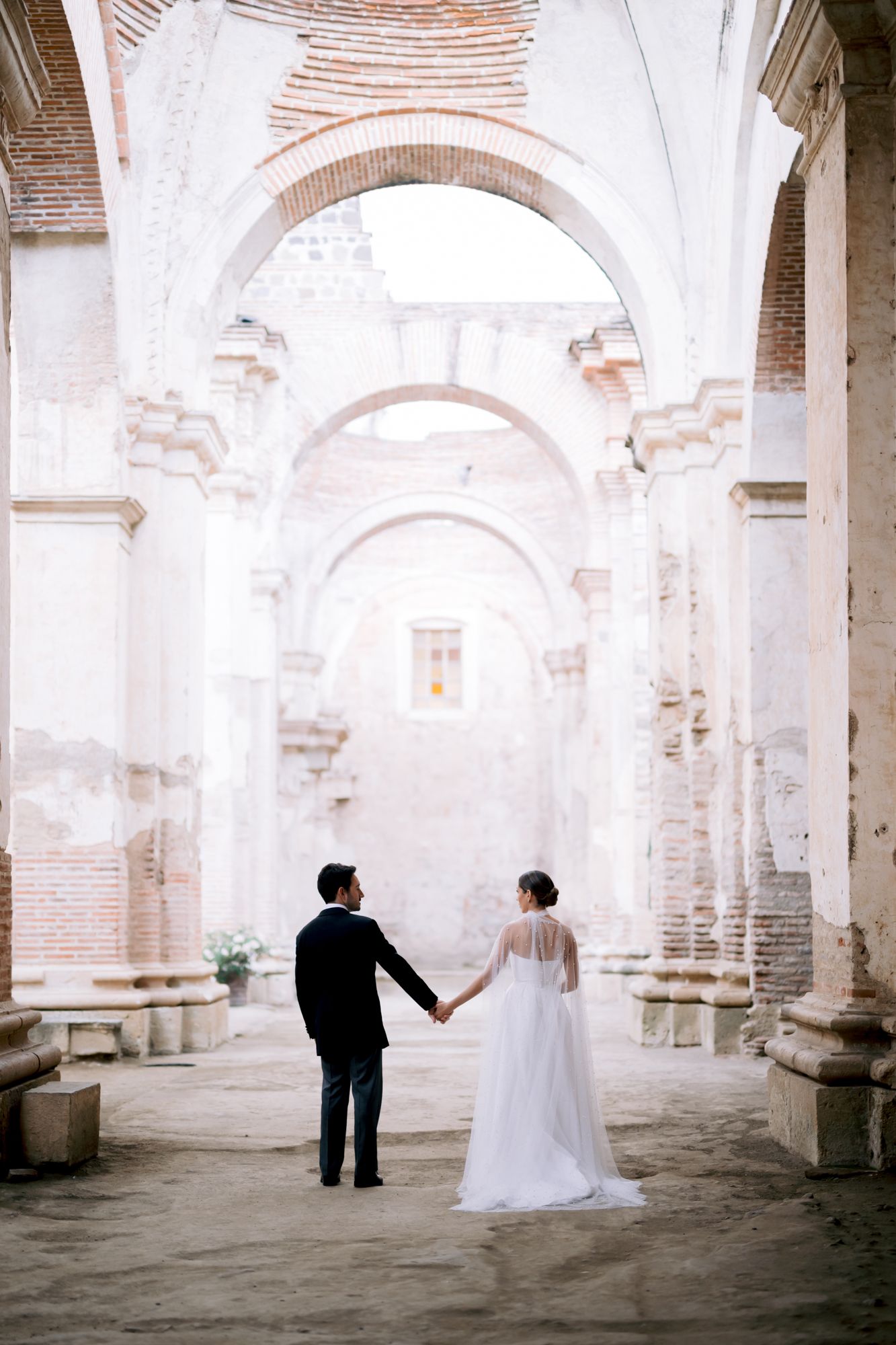 Bride and groom hold hands while walking through beautiful stone archways in historic building.