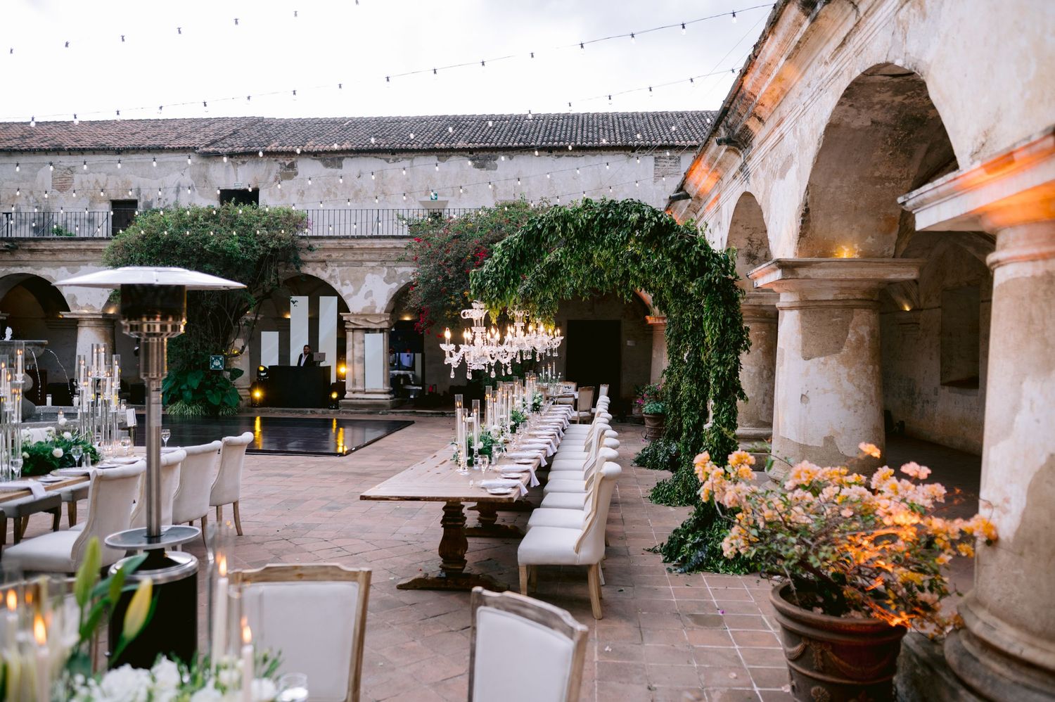 Long dining tables arranged in outdoor courtyard with string lights and chandeliers.