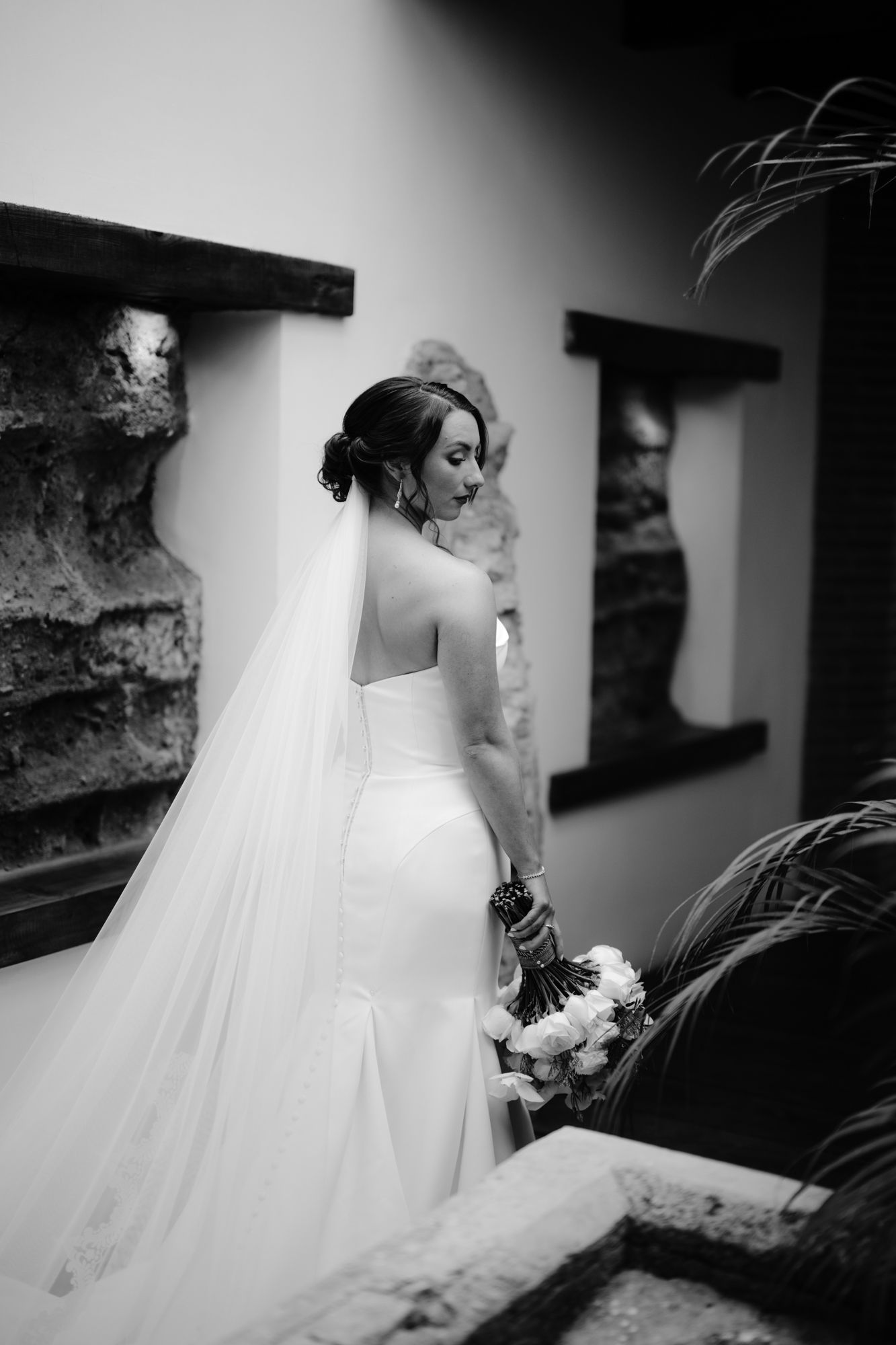 A black and white photo of a bride with flowing veil looking over her shoulder.