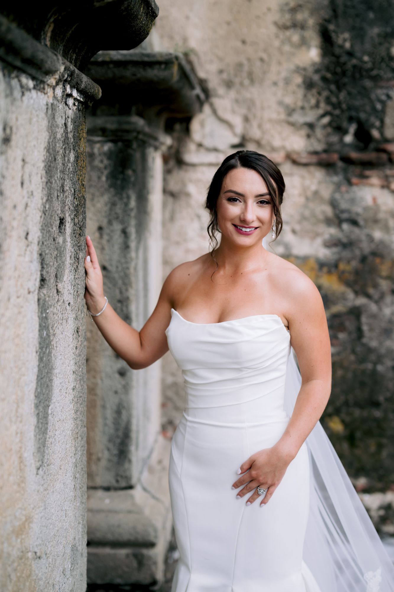 A person in a strapless white wedding dress poses against an old stone wall with a joyful expression.