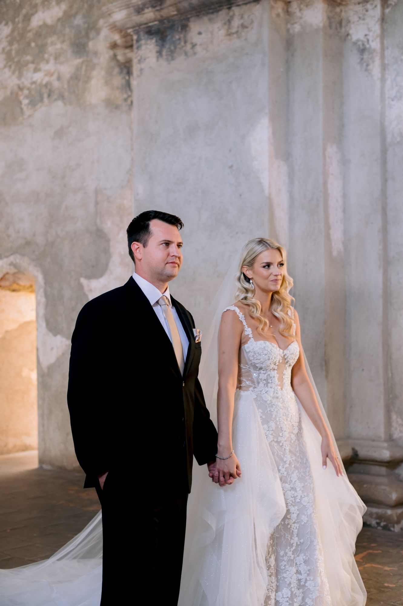 Bride in lace gown and groom in black tuxedo pose elegantly in historic stone building.