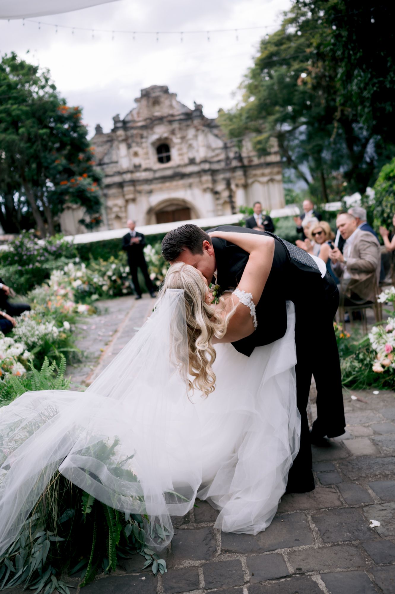 Romantic dip pose during wedding celebration with historic church and garden setting.