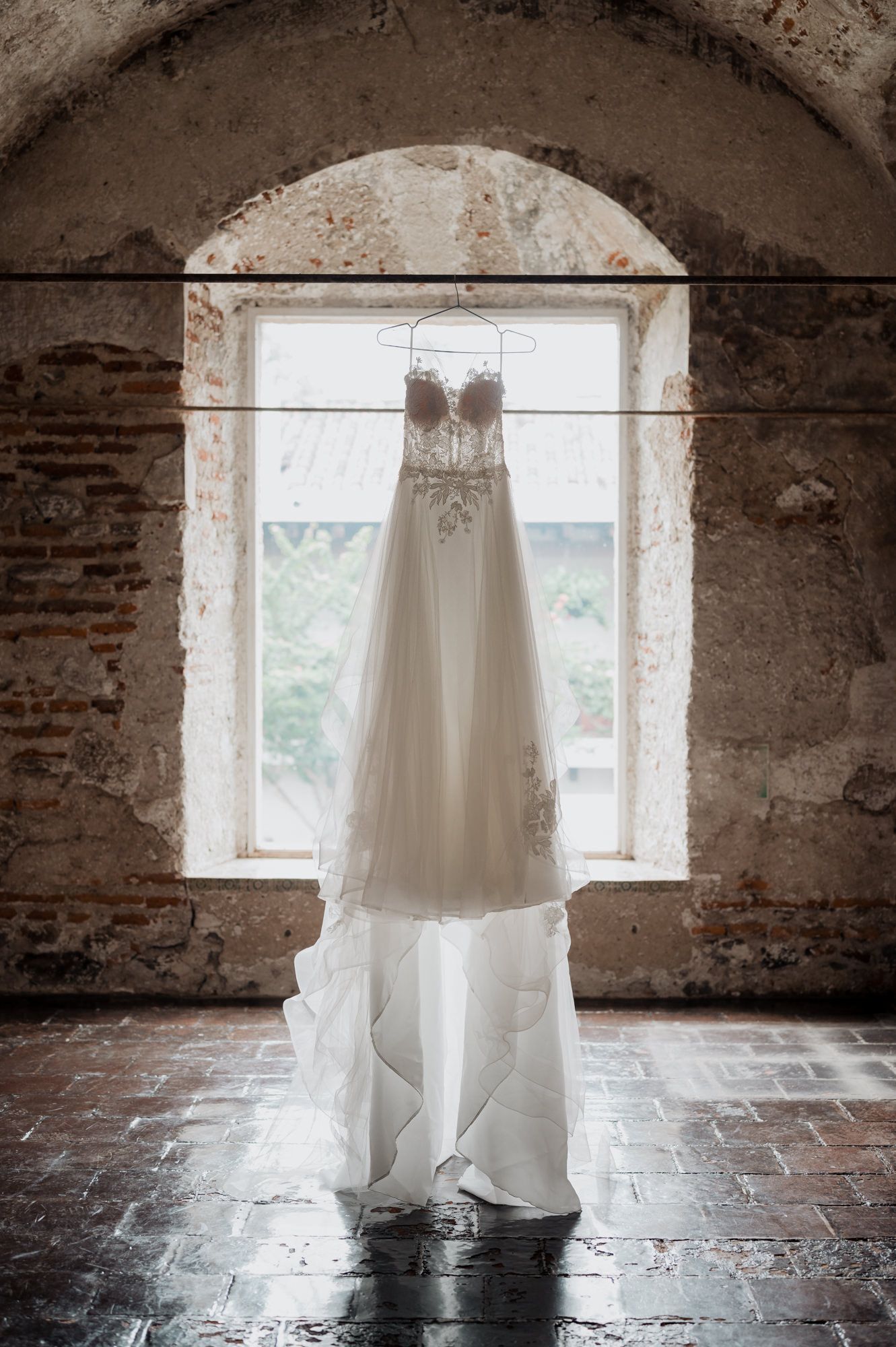 A white wedding dress hangs elegantly in a rustic stone window arch creating a dramatic silhouette.